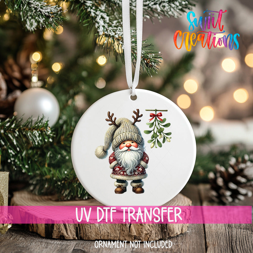 A white ornament with a gnome and mistletoe hangs on a Christmas tree, surrounded by pine branches and twinkling lights.