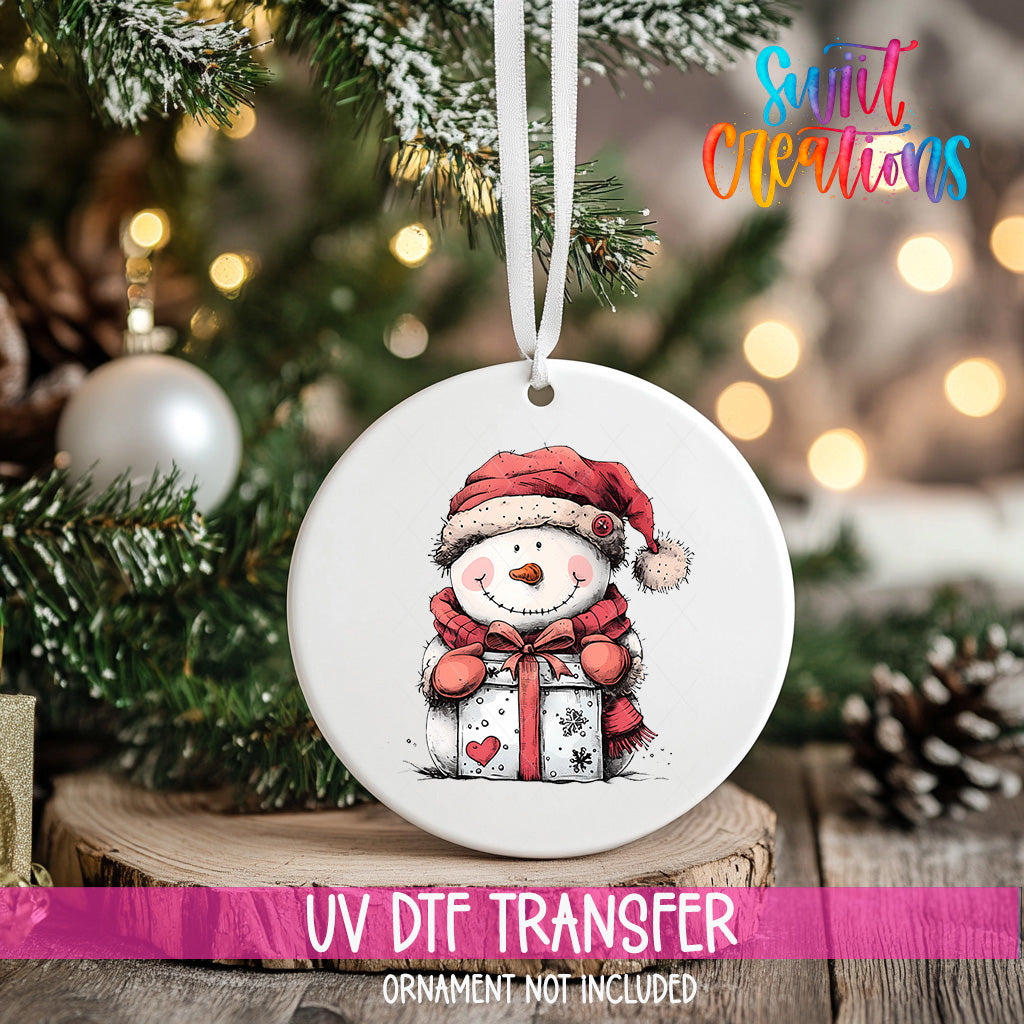 A white ceramic ornament with a snowman and a gift is hanging on a Christmas tree, with a pink ribbon below it.