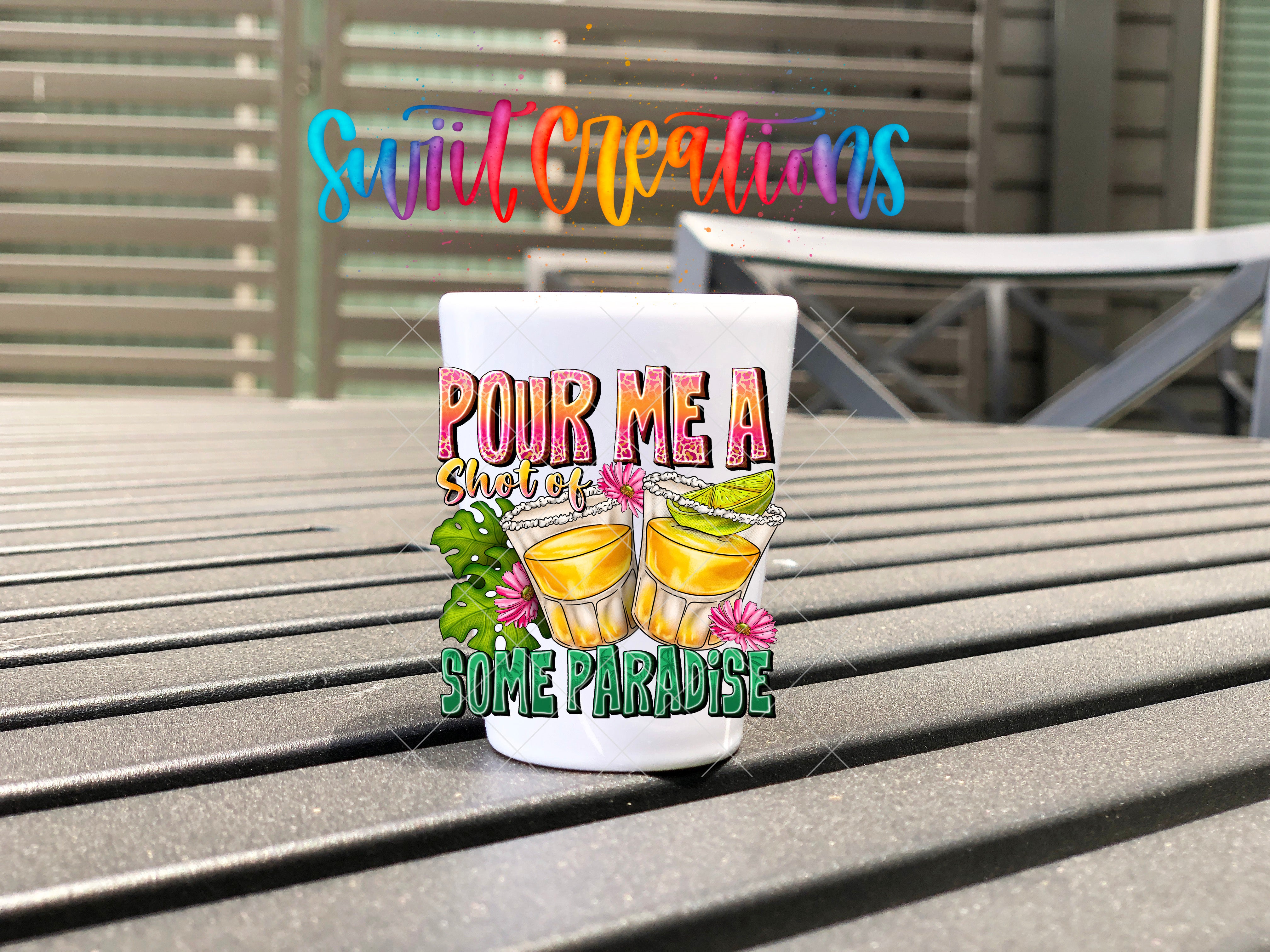 A colorful shot glass with the text "POUR ME A SHIT" and "SOME PARADISE" printed on it, placed on a wooden surface.