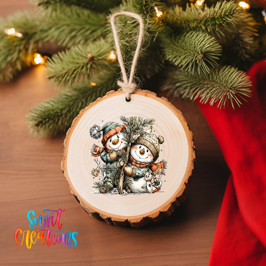 A wooden ornament featuring two snowmen is hanging on a Christmas tree, surrounded by pine branches and twinkling lights.