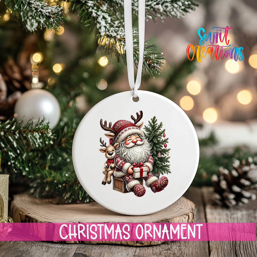 A white Christmas ornament featuring a Santa Claus figure holding a reindeer and a present, hanging on a tree branch with a blurred background of lights and greenery.