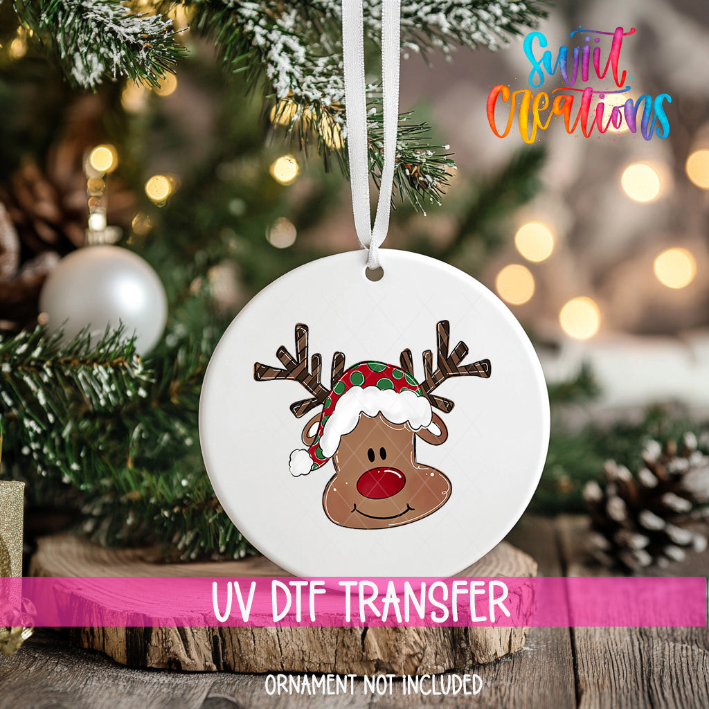 A white ornament with a cartoon reindeer wearing a Santa hat is hanging on a Christmas tree.