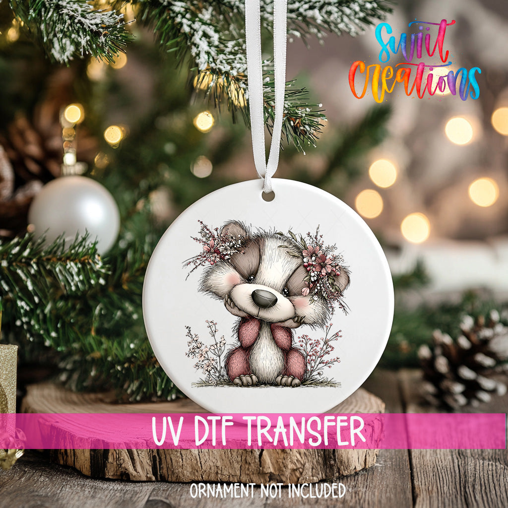 A white ceramic ornament with a cute cartoon bear design hangs on a Christmas tree, surrounded by pine branches and other holiday decorations.