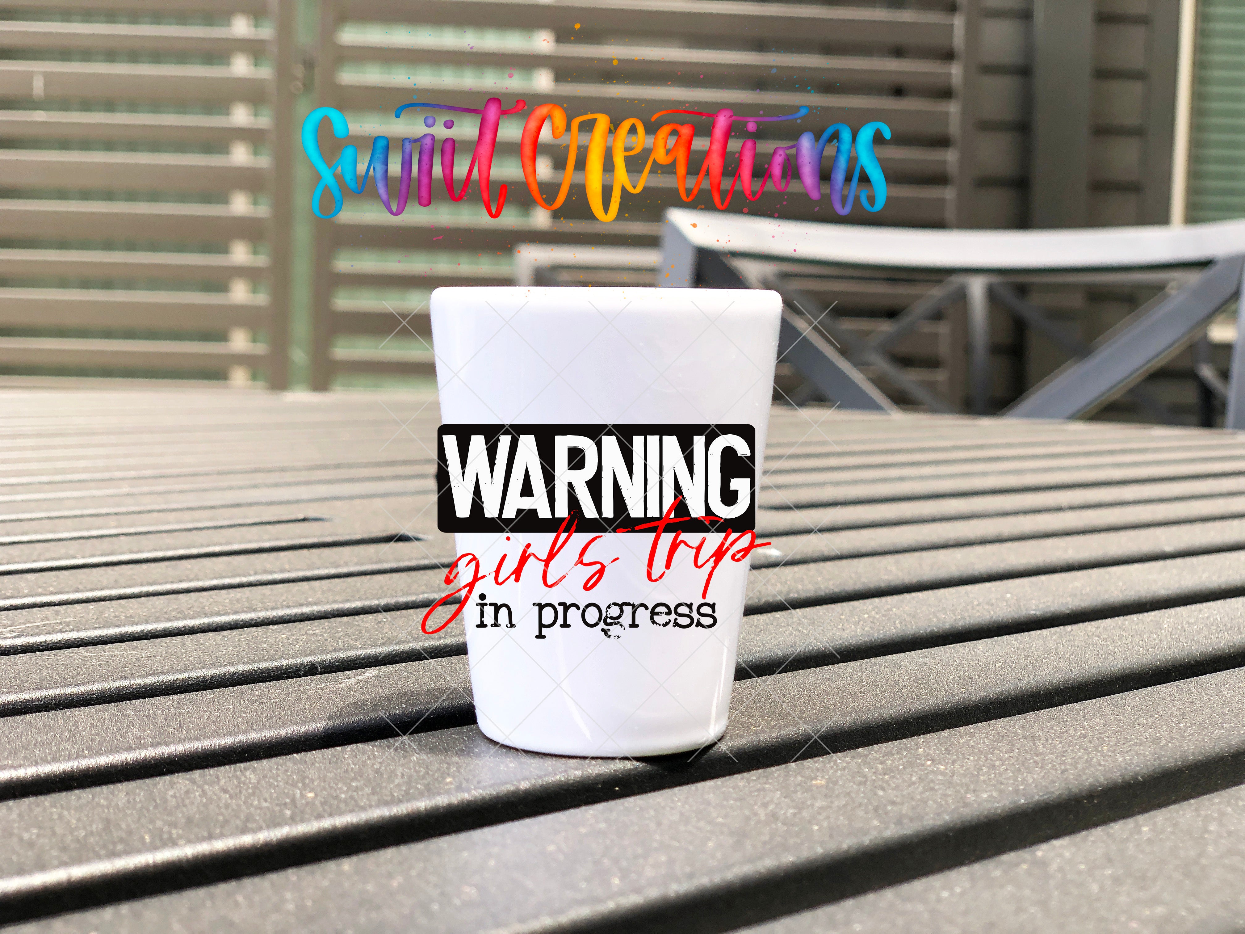 A white shot glass with the text "WARNING girls trip in progress" printed on it, placed on a wooden surface.