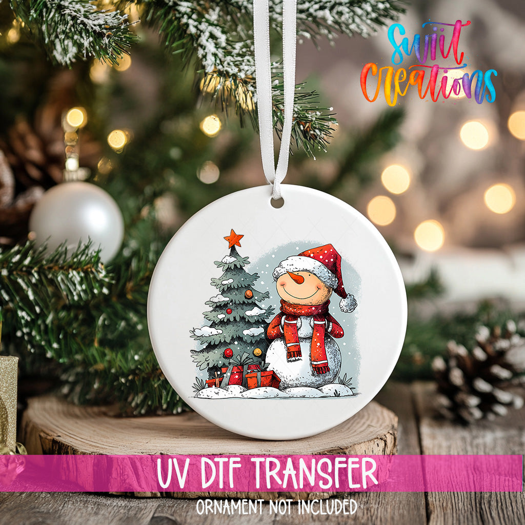 A white Christmas ornament with a snowman and a Christmas tree, hanging on a tree branch with a ribbon.
