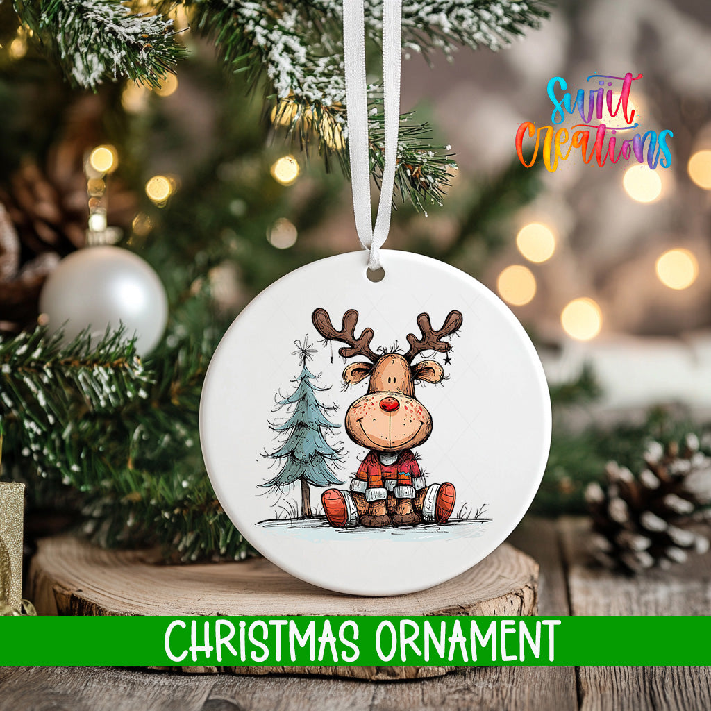 A white Christmas ornament featuring a cartoon reindeer sitting on a pine tree, with a blurred background of a decorated Christmas tree and twinkling lights.
