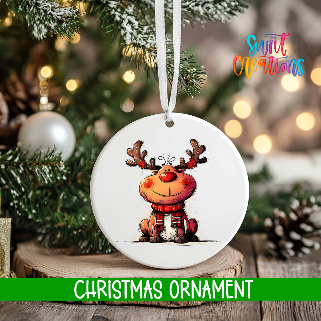A white Christmas ornament featuring a cartoon reindeer is hanging on a tree branch, with a blurred background of lights and greenery.