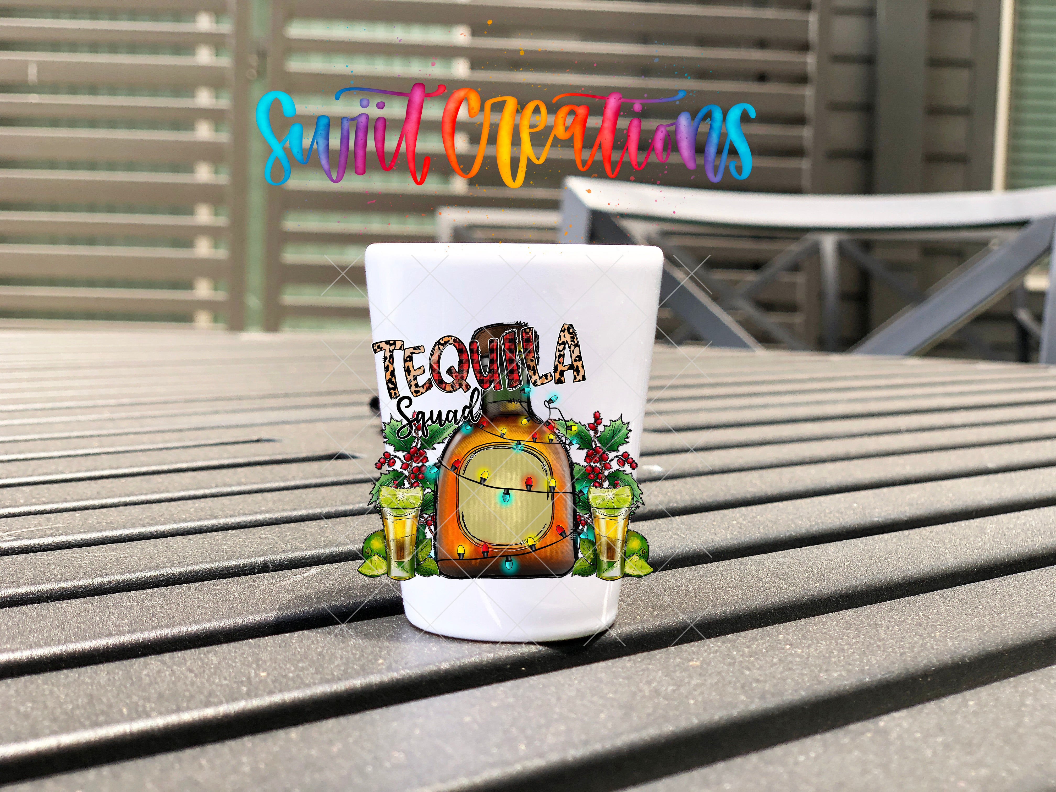 A white shot glass with a colorful design featuring a tequila bottle, lime slices, and other festive elements, placed on a wooden surface.