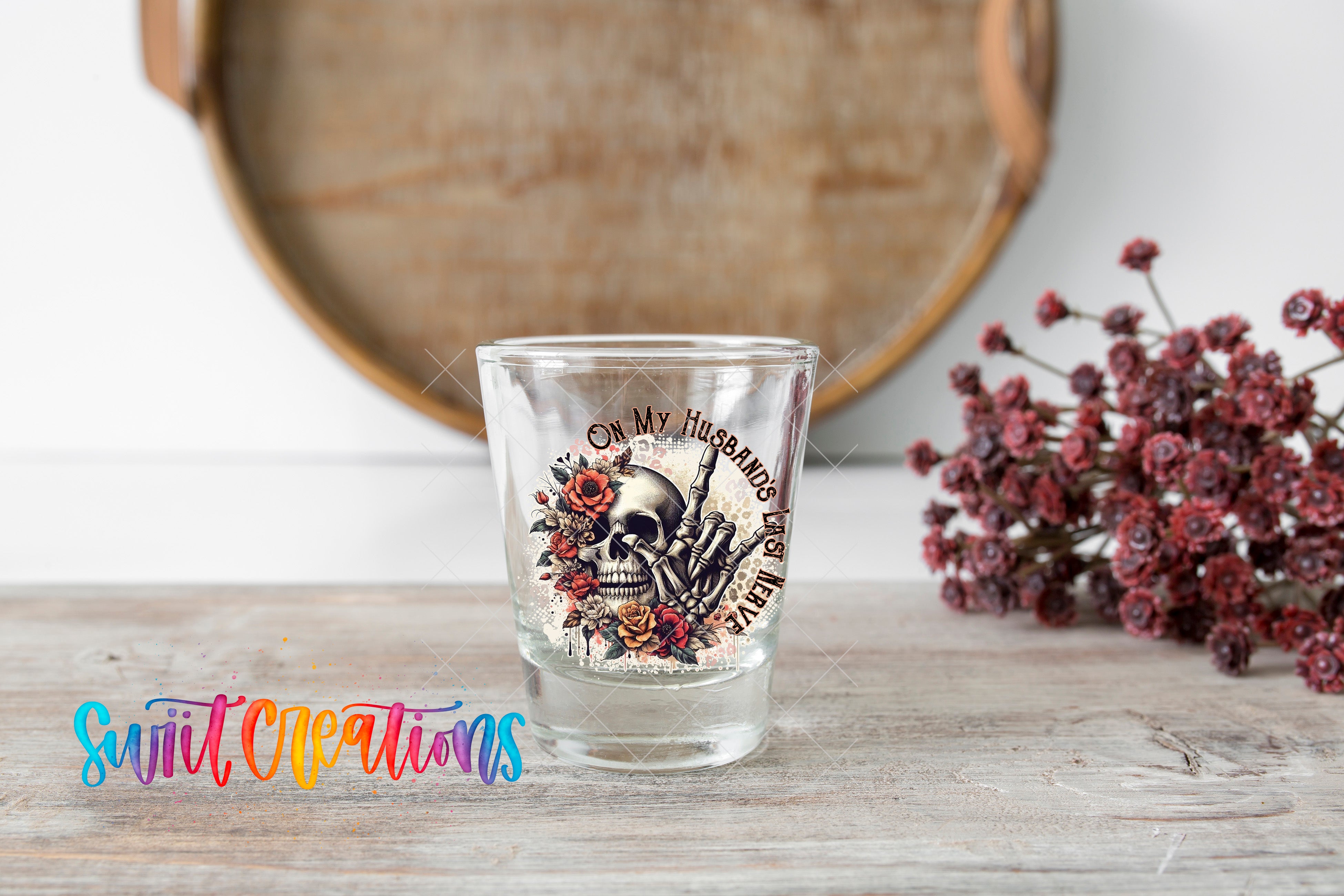A shot glass with a skull design and floral elements is placed on a wooden surface, with a wooden tray and some flowers in the background.
