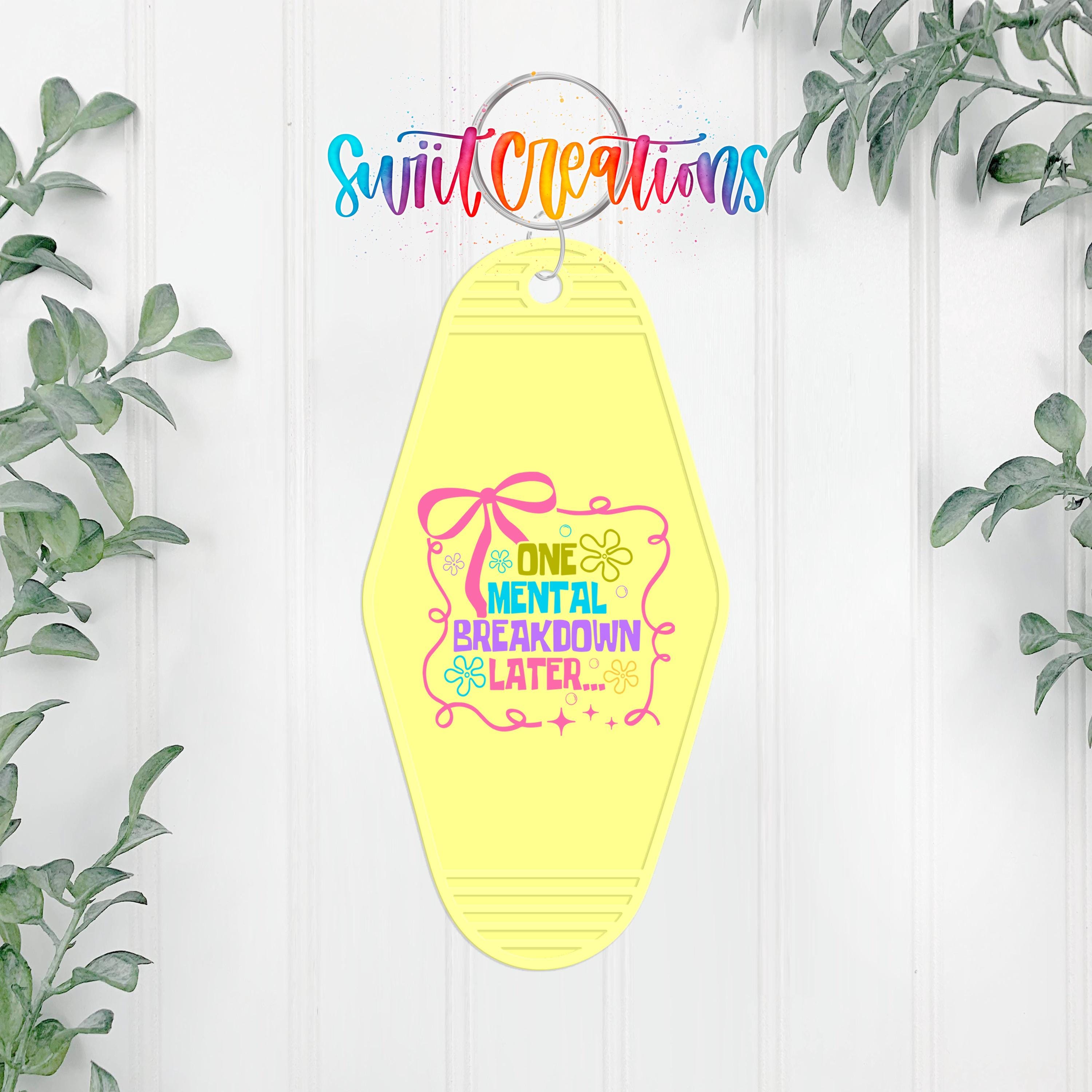 A yellow keychain with a colorful graphic and text, hanging on a white wall with greenery in the background.