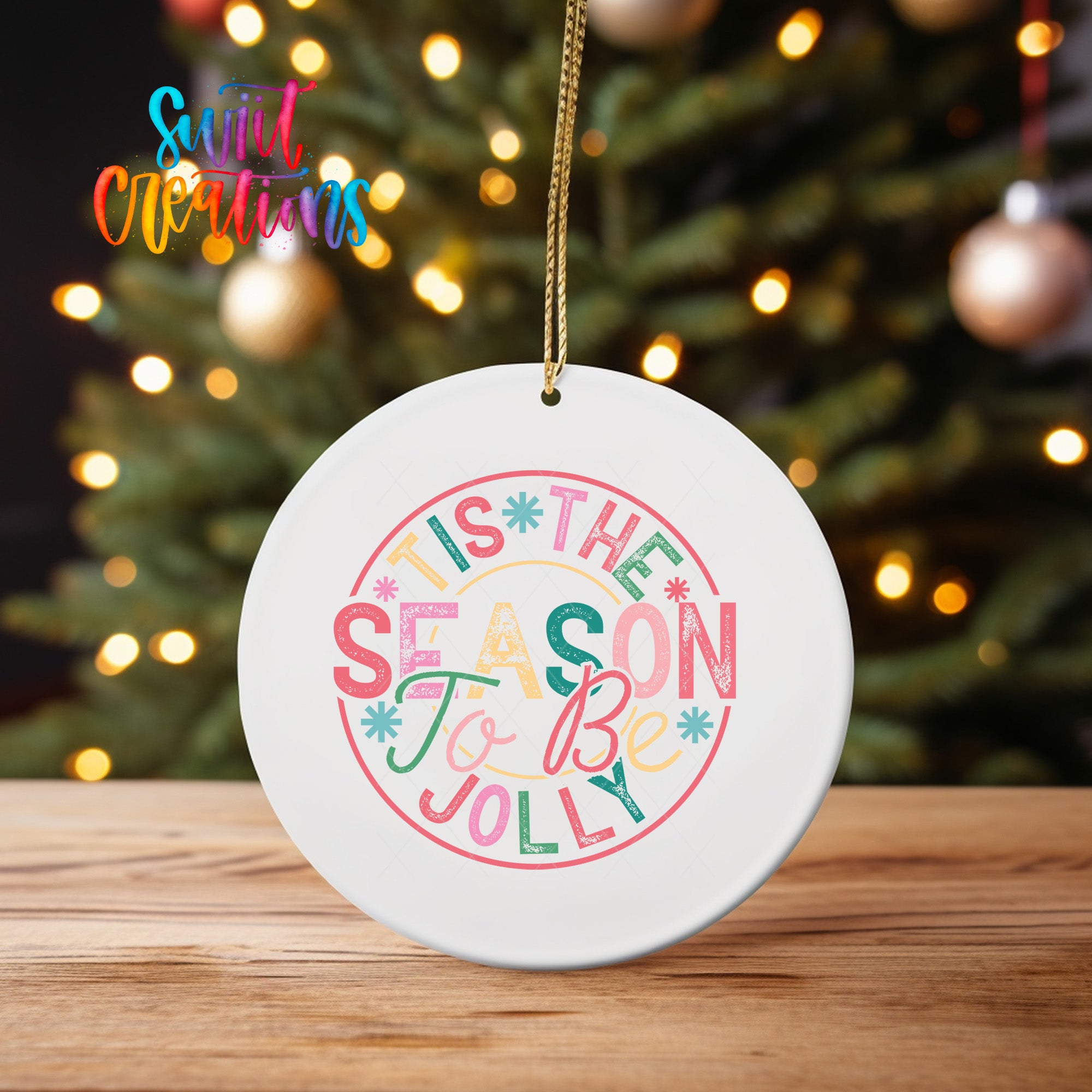 A white ceramic ornament with a colorful design hangs on a wooden surface, with a blurred background of a Christmas tree and twinkling lights.