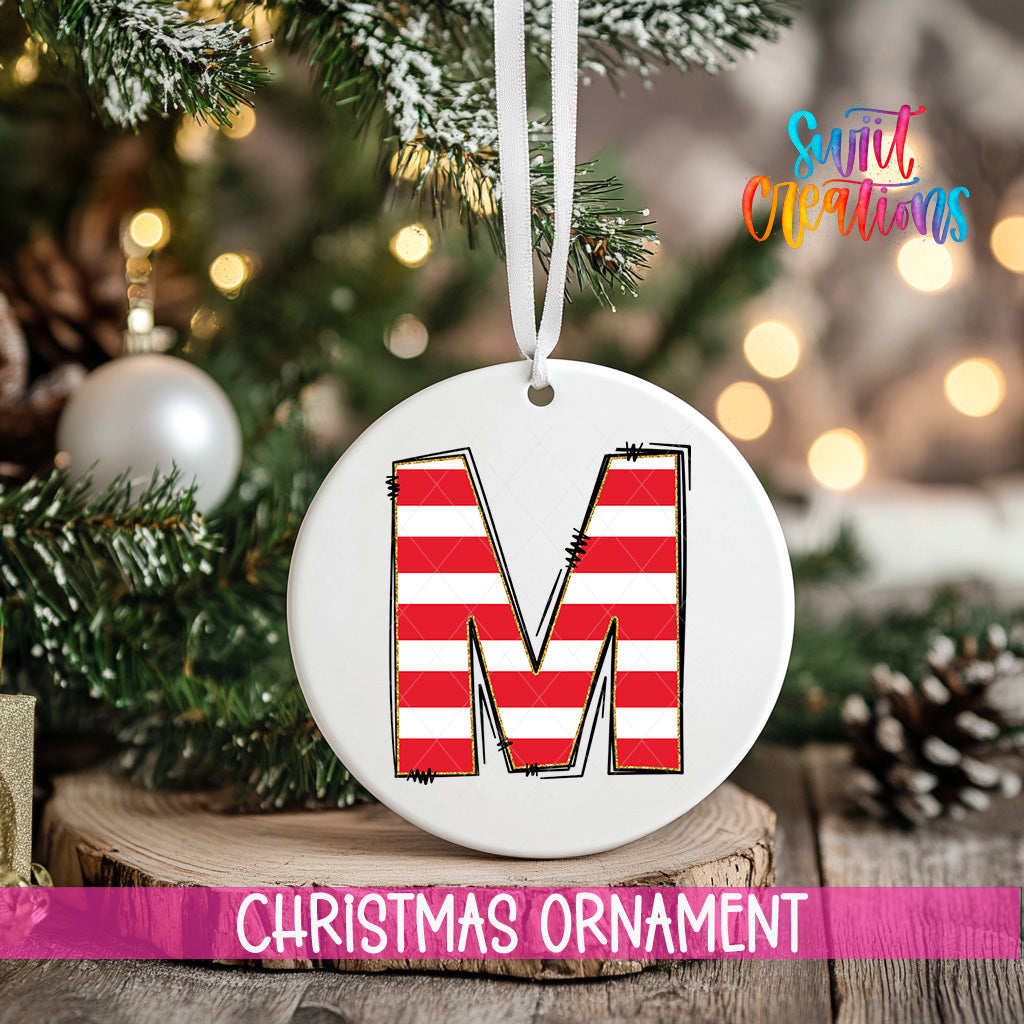 A white Christmas ornament with a red and white striped design, featuring the letter "M" in the center, is hanging from a tree branch.