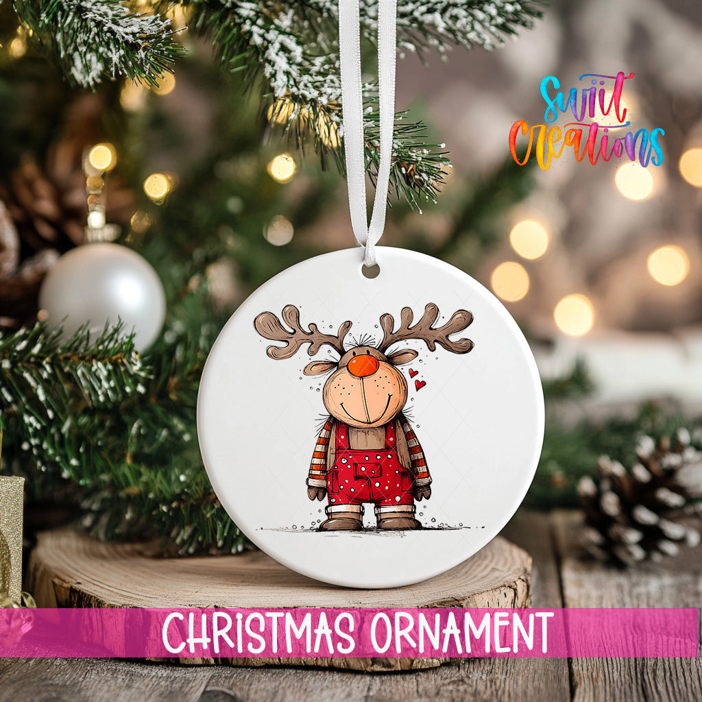 A white Christmas ornament featuring a cartoon reindeer is hanging from a tree branch, with a blurred background of lights and greenery.
