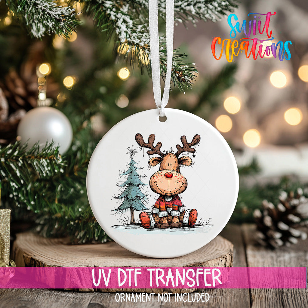 A white ceramic ornament with a cartoon reindeer sitting on a pine tree, surrounded by Christmas decorations and lights.