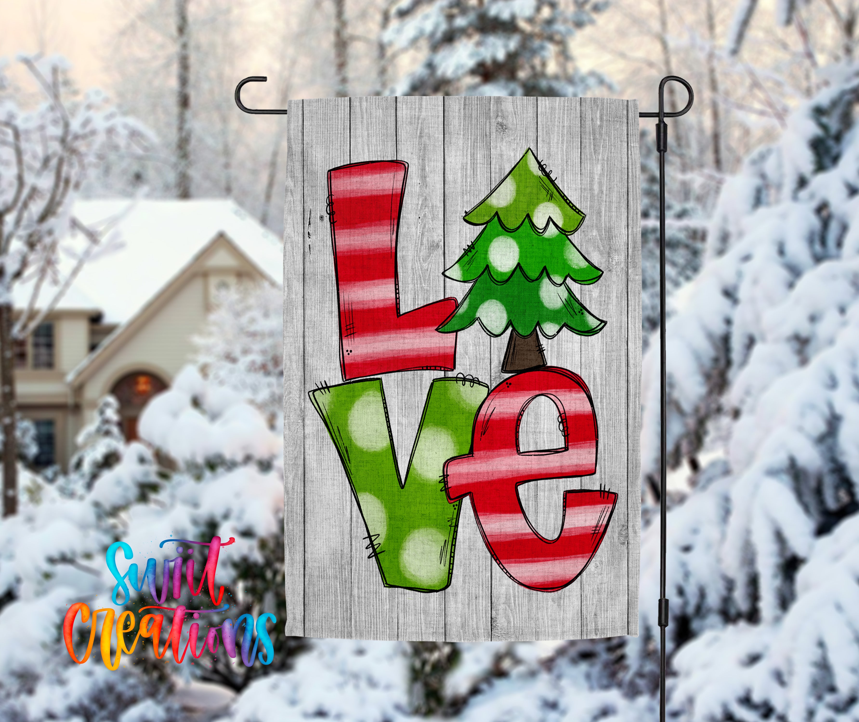A wooden flag with the word "LOVE" written on it, featuring a Christmas tree design. The flag is hanging in front of a snowy landscape with a house and trees in the background.