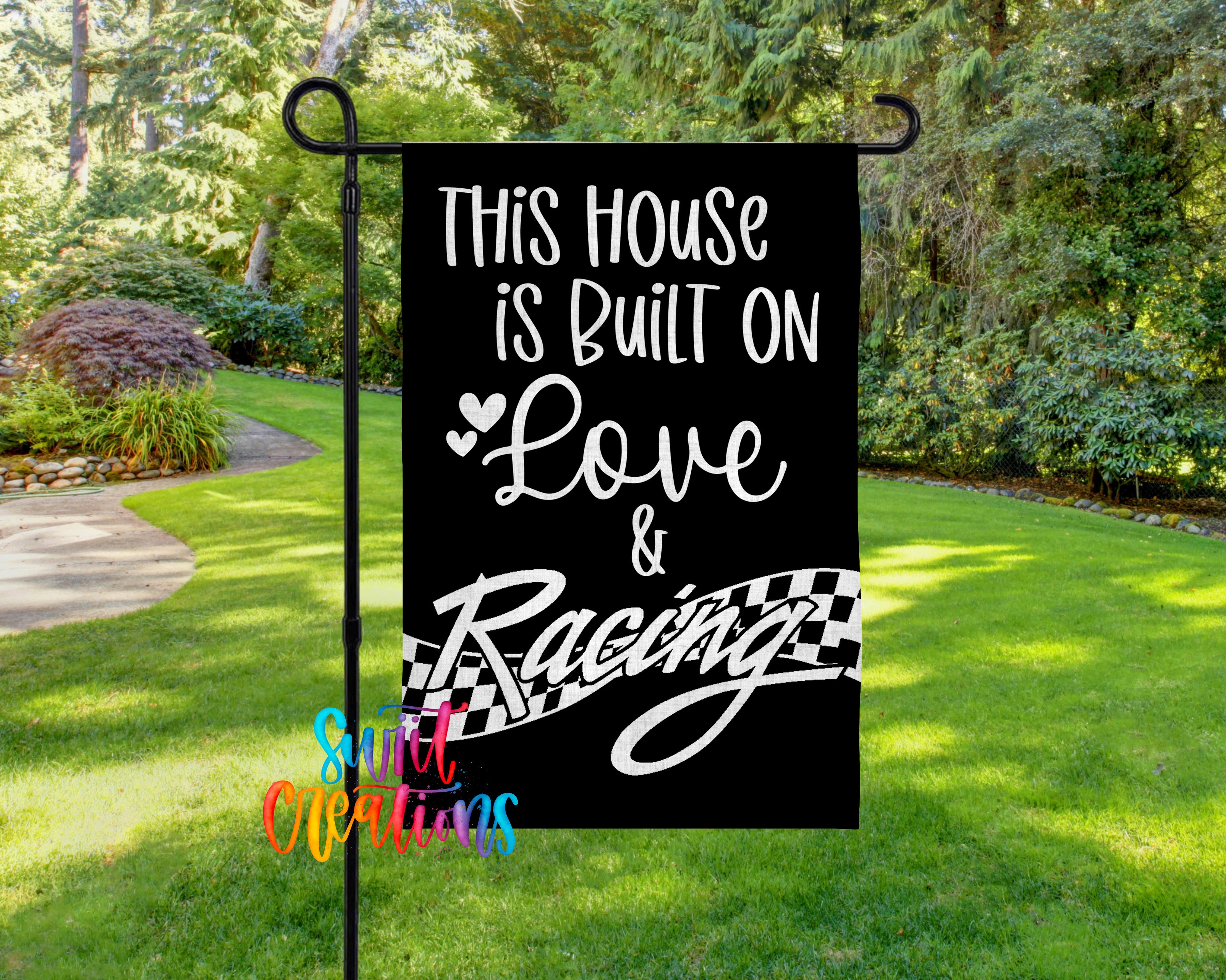 a black and white sign with the text "This house is built on love & racing" displayed on it, hanging in a grassy area surrounded by trees.
