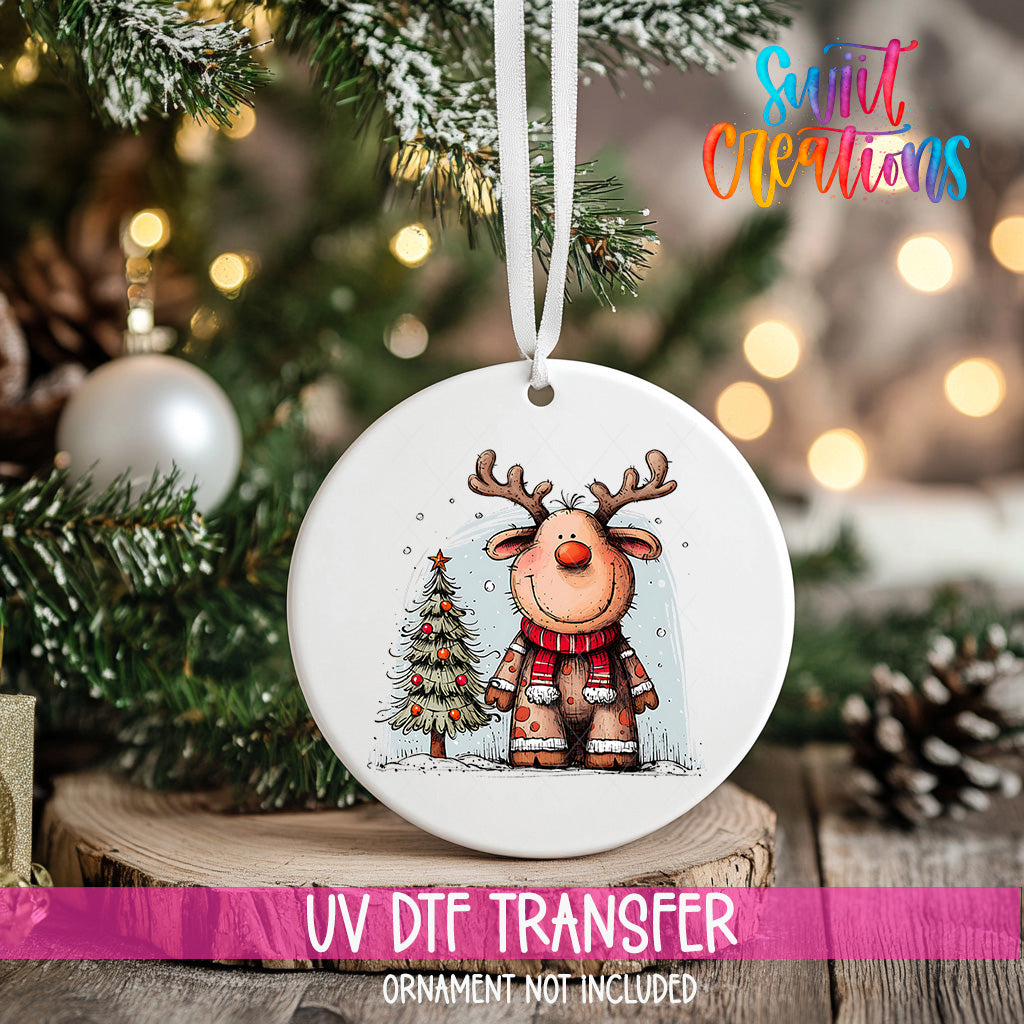 A white ornament with a cartoon reindeer and a Christmas tree is hanging on a tree branch, with a blurred background of lights and greenery.
