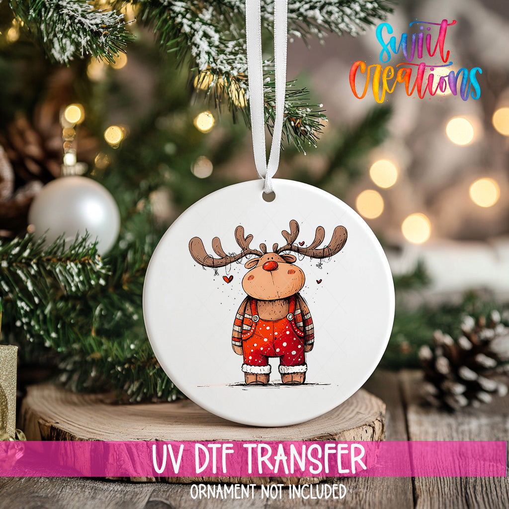 A white ornament with a cartoon reindeer design hangs on a Christmas tree, surrounded by pine branches and twinkling lights.