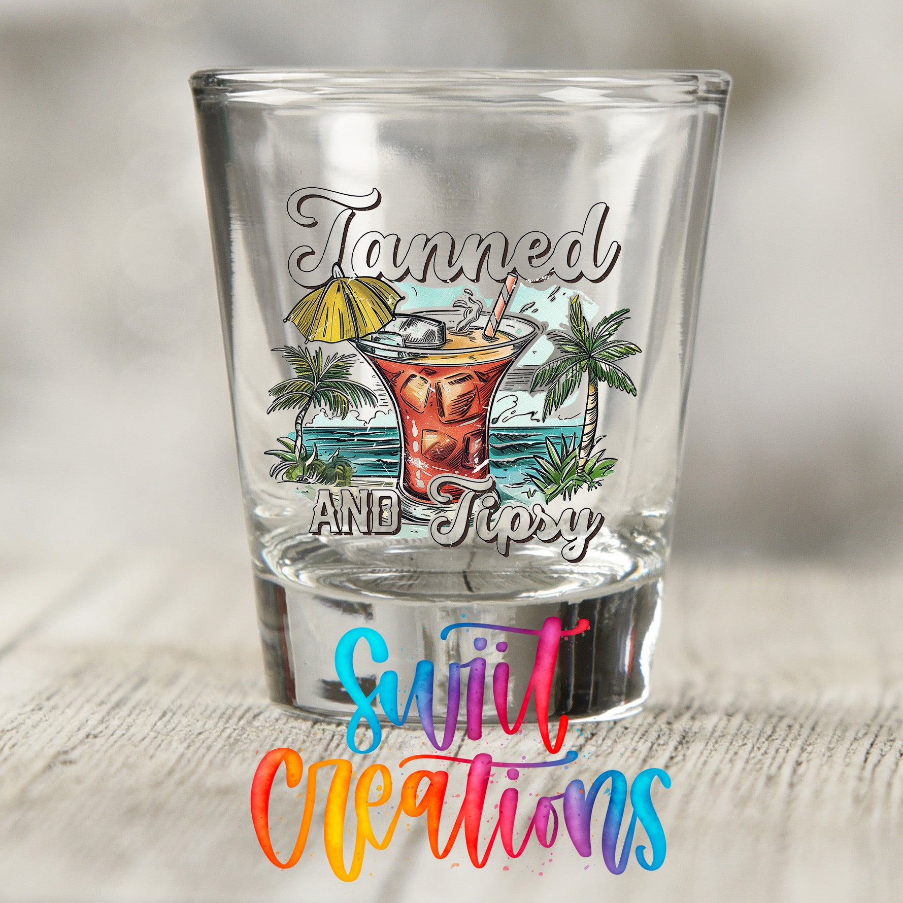A shot glass with a colorful design featuring a tropical drink and palm trees, along with the text "Tanned AND Tipsy" and "Sweet Creations" printed on it.