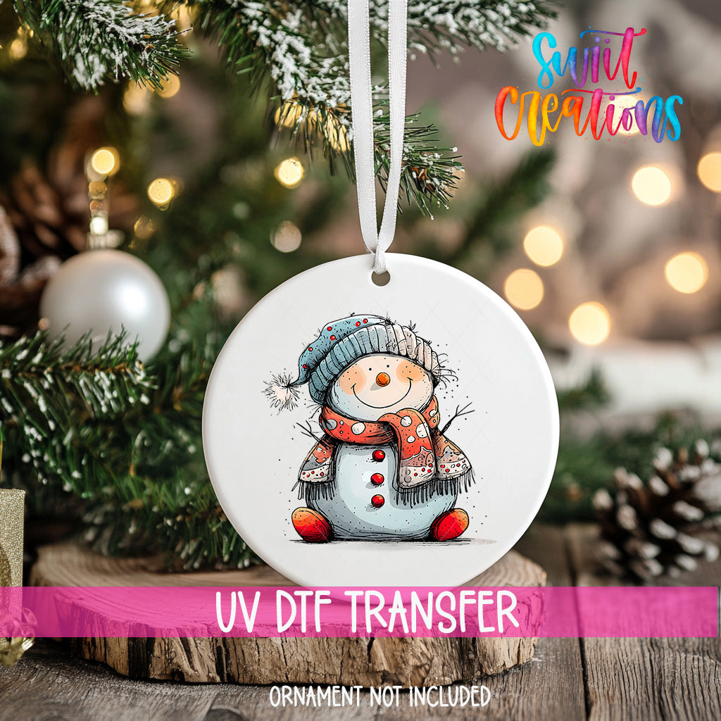 A white ornament with a snowman design hangs on a Christmas tree, surrounded by pine branches and twinkling lights.