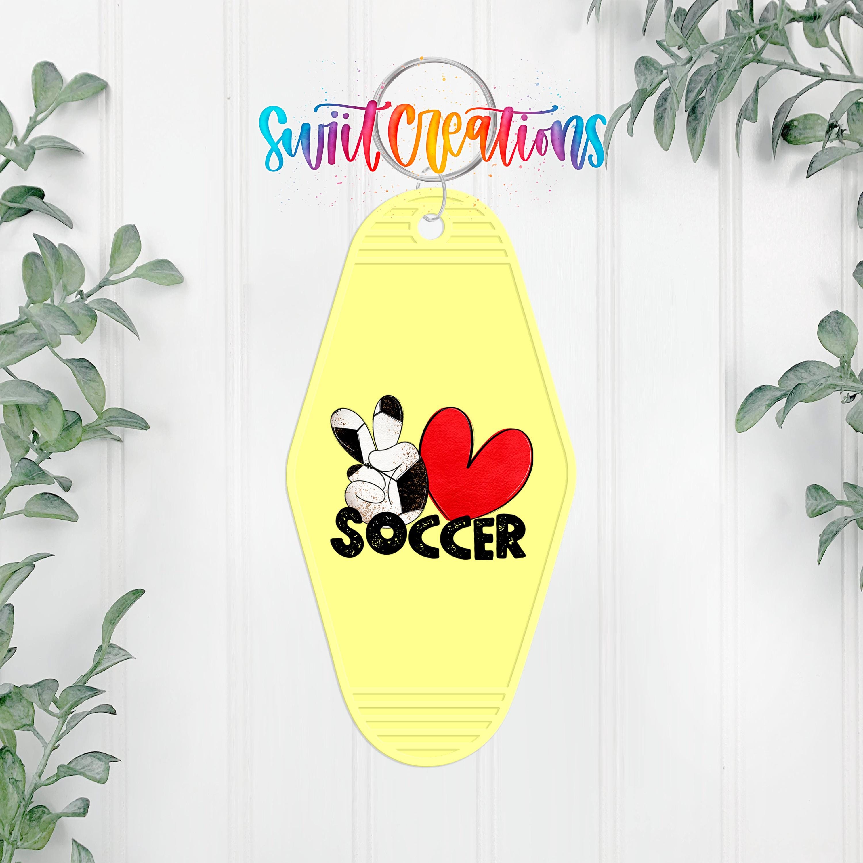 A yellow keychain with a soccer-themed design and the word "SOCCER" printed on it, hanging on a white wall with greenery in the background.