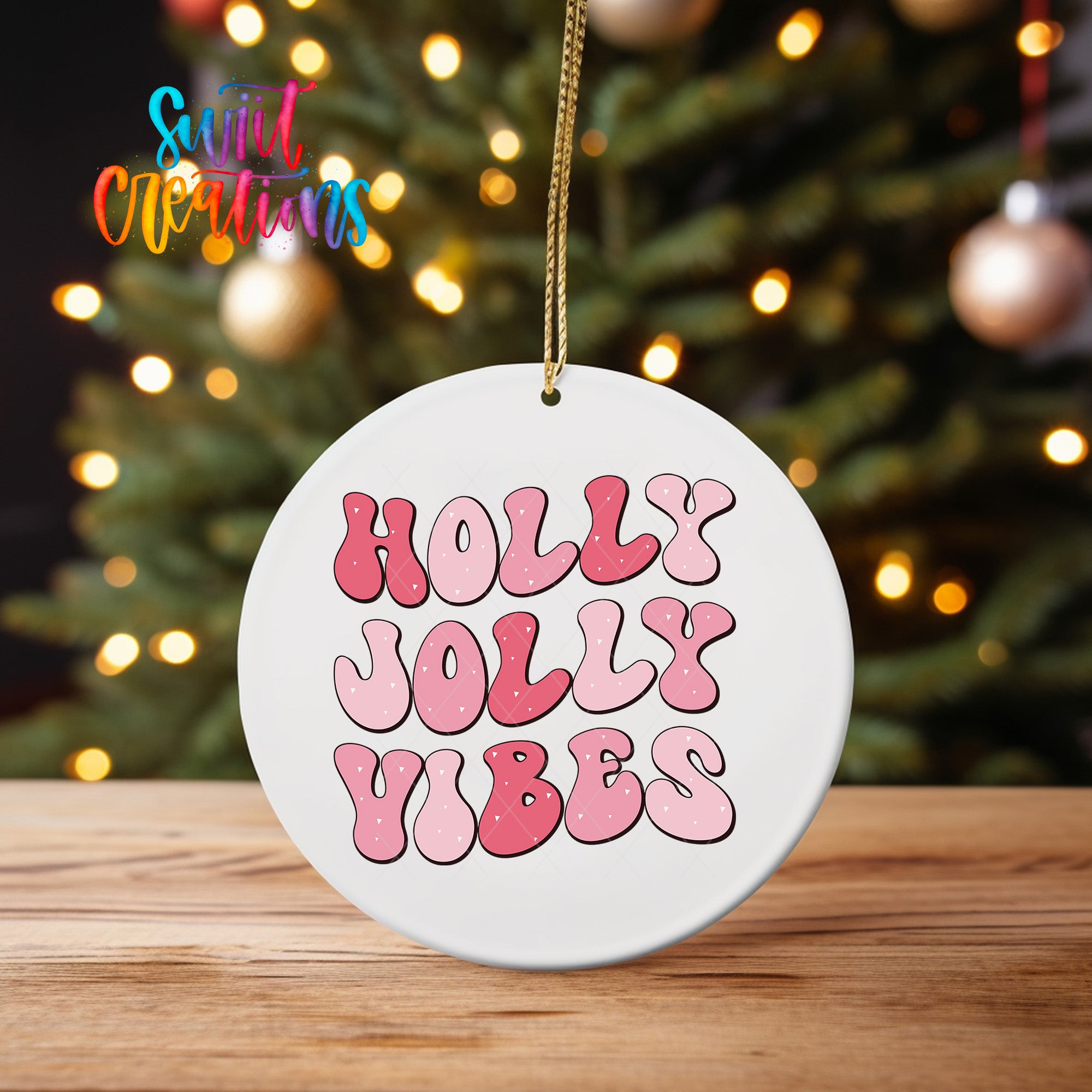 A white ornament with the text "Holly Jolly Vibes" in pink and red letters is hanging from a string in front of a blurred Christmas tree.