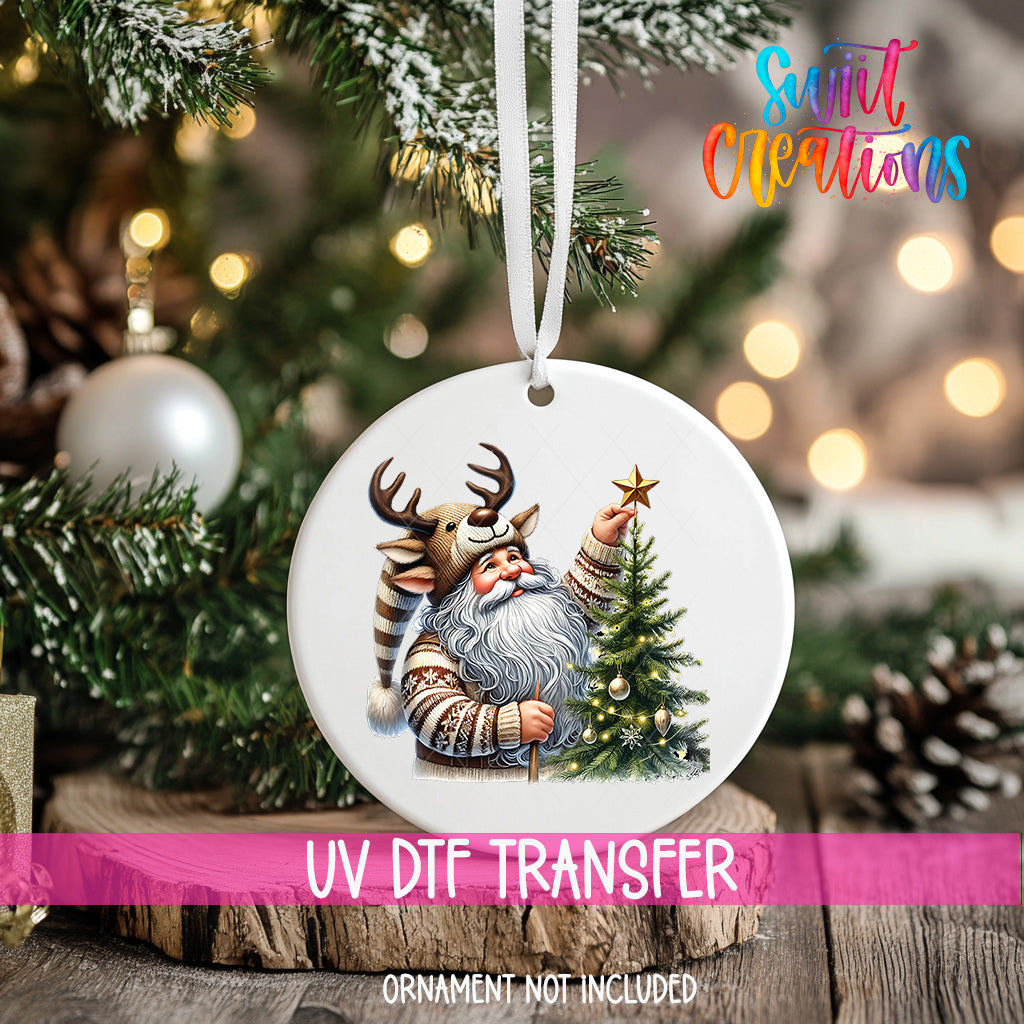 A white ornament with a Santa Claus figure and a Christmas tree is hanging on a tree branch, with a pink ribbon around it.