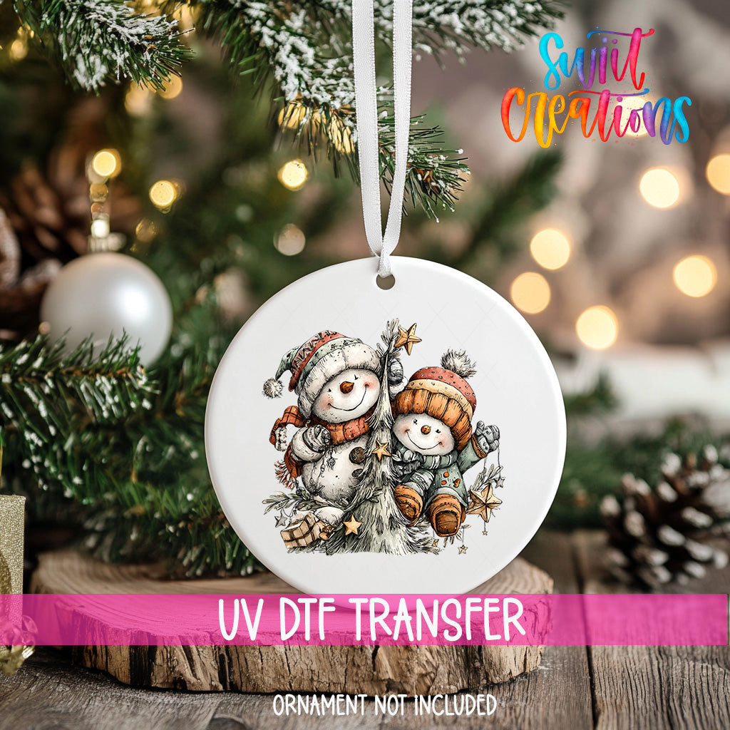 A white ornament with a snowman and star on a Christmas tree, with a pink ribbon around it.