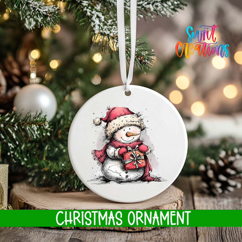A white Christmas ornament featuring a snowman holding a gift, hanging on a tree branch with a blurred background of lights and greenery.