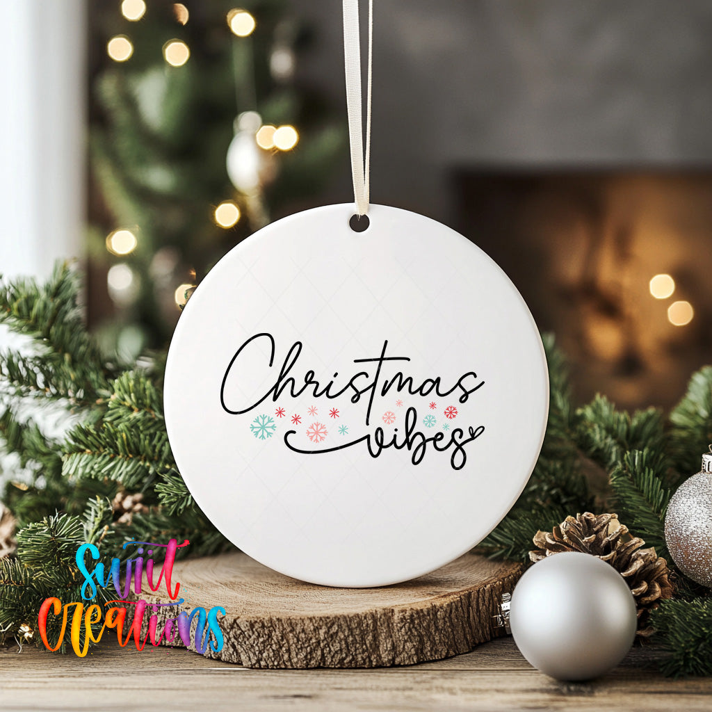 A white Christmas ornament with the text "Christmas vibes" written on it, hanging from a string. The ornament is surrounded by pine branches and other holiday decorations, creating a festive atmosphere.