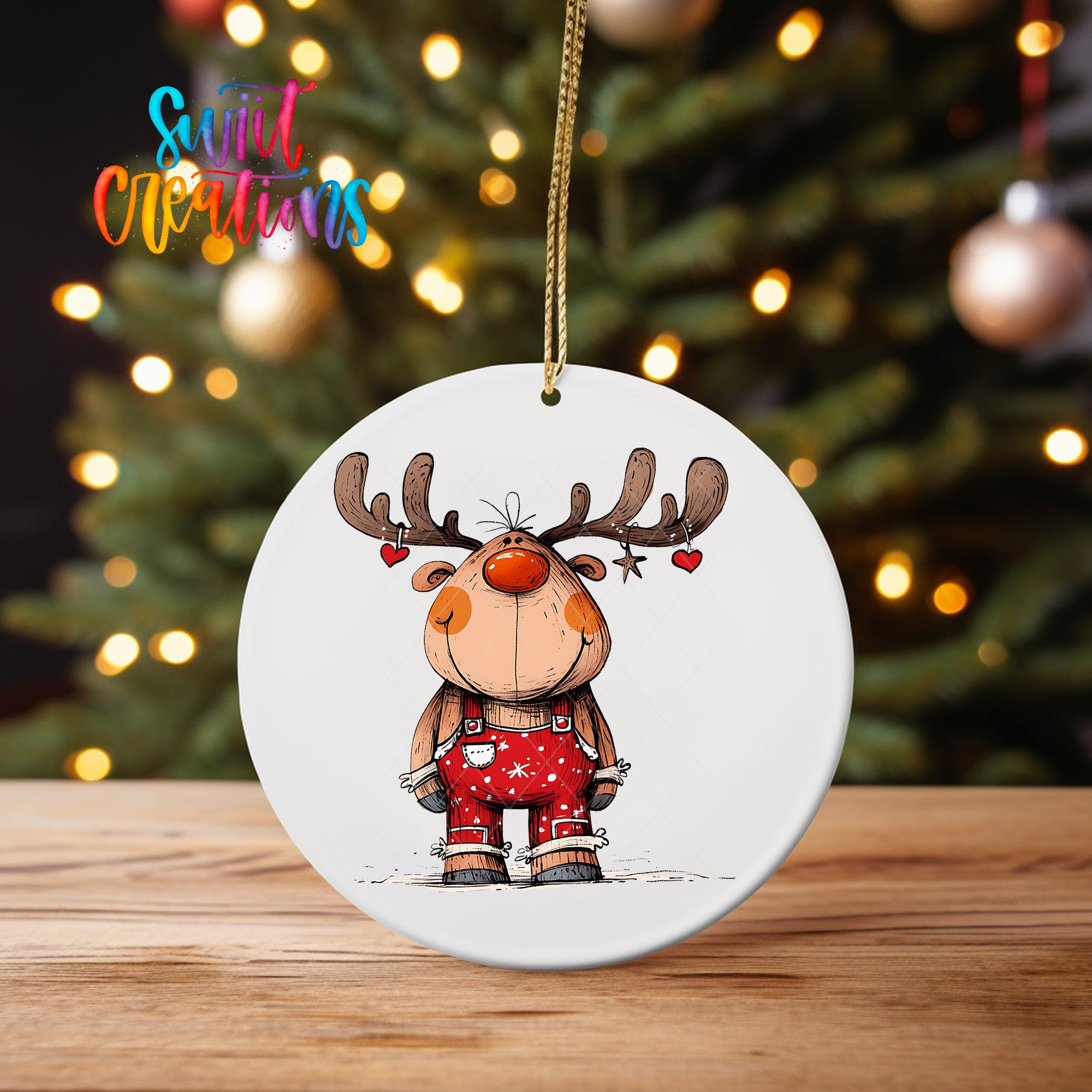 A white ornament featuring a cartoon reindeer with antlers and a red nose, standing on a wooden surface in front of a blurred Christmas tree with twinkling lights.