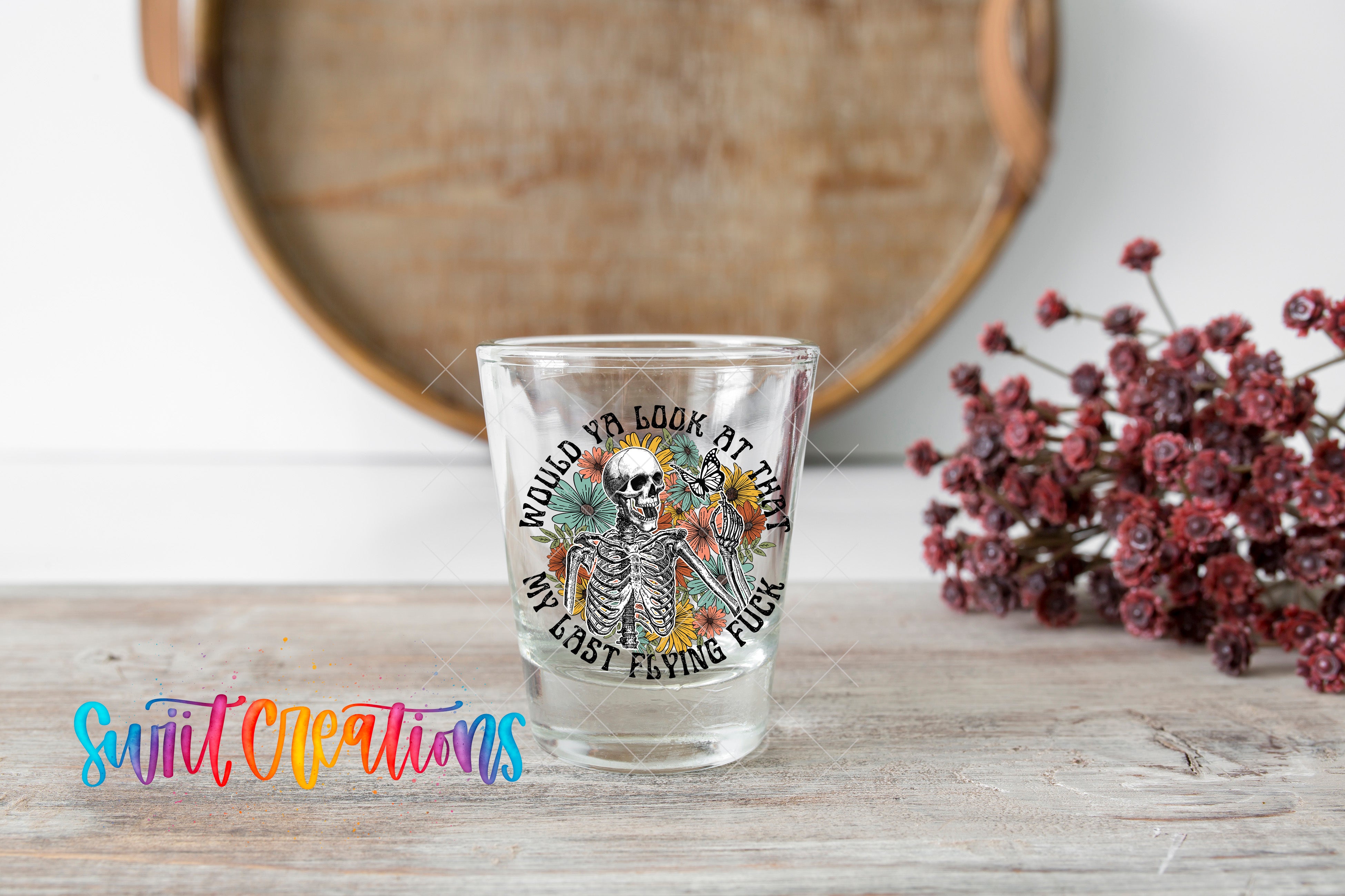 A shot glass with a skeleton design and text on it, placed on a wooden surface next to a bunch of red flowers.