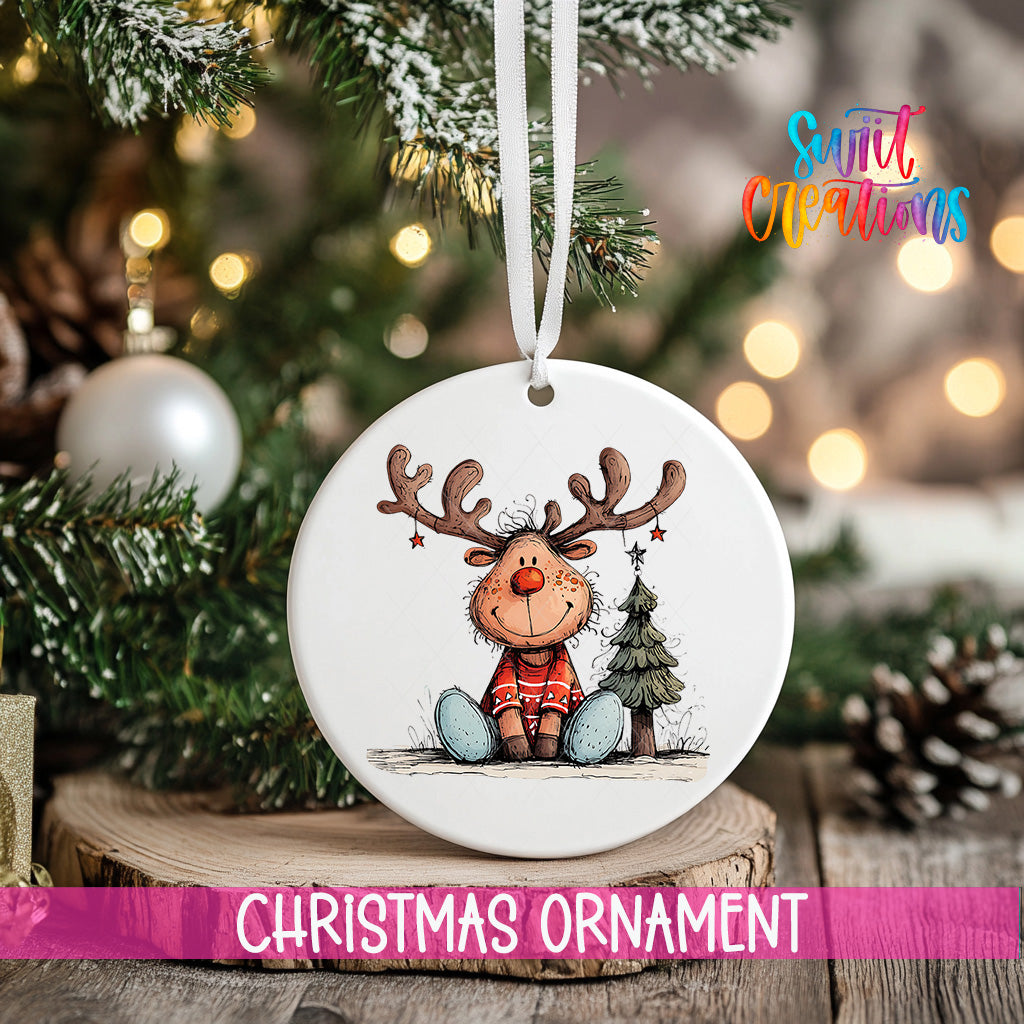 A white Christmas ornament featuring a cartoon reindeer sitting on a Christmas tree, with a tree branch and pine cones in the background.
