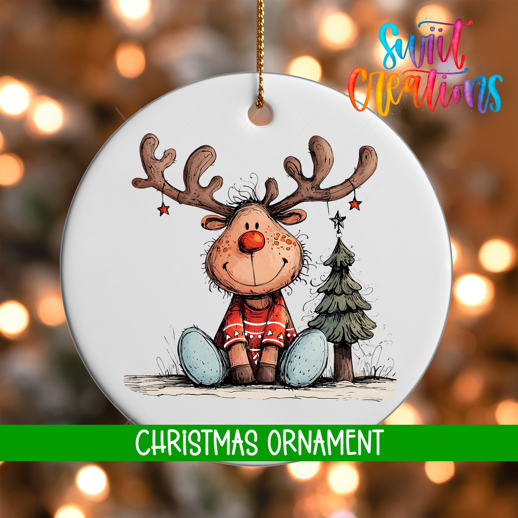 A white Christmas ornament featuring a smiling reindeer with antlers and a star, sitting next to a small Christmas tree.