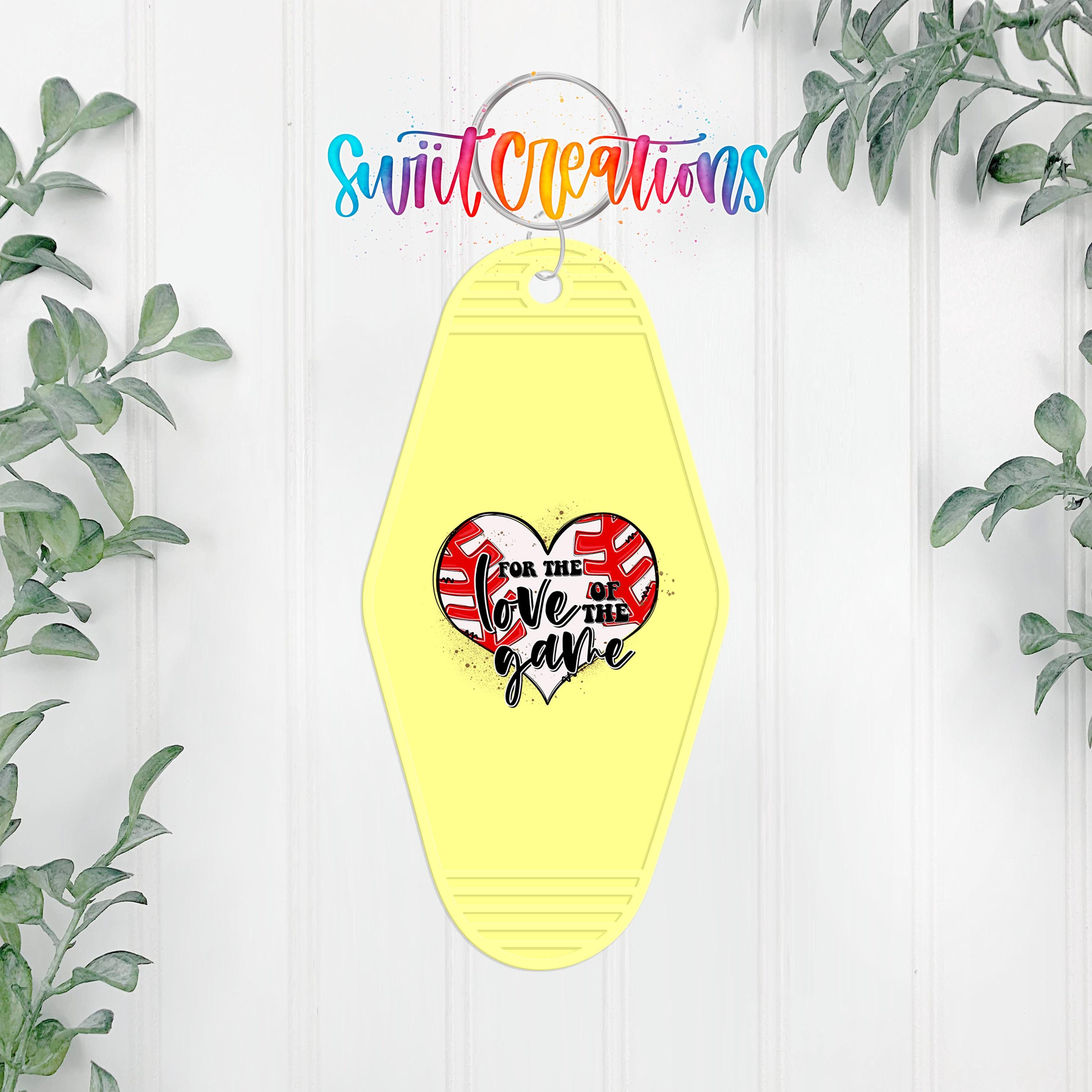 A yellow keychain with a heart-shaped design and text on it, hanging on a white wall with greenery in the background.