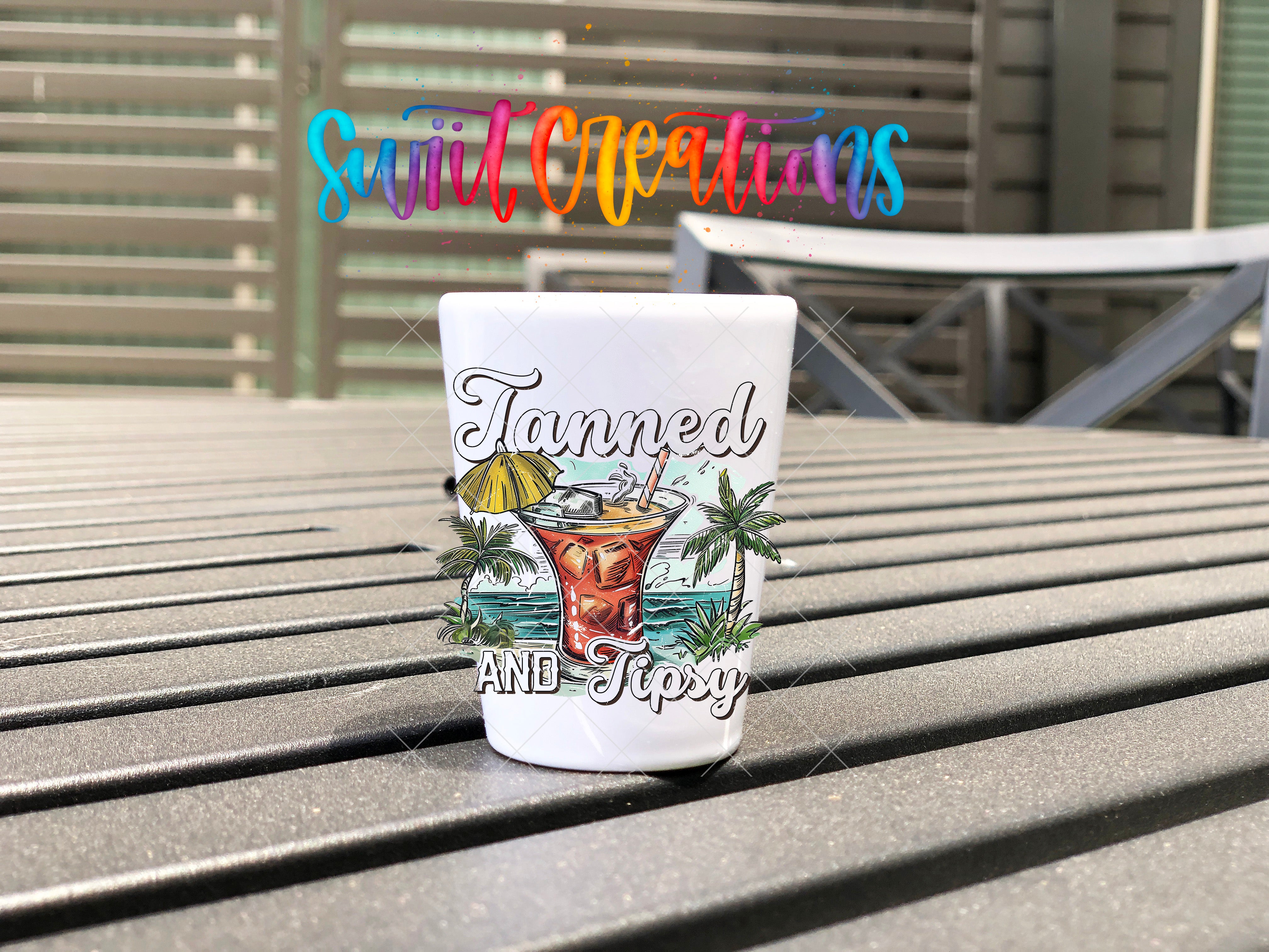 A white shot glass with a colorful design and text on it, placed on a wooden surface.