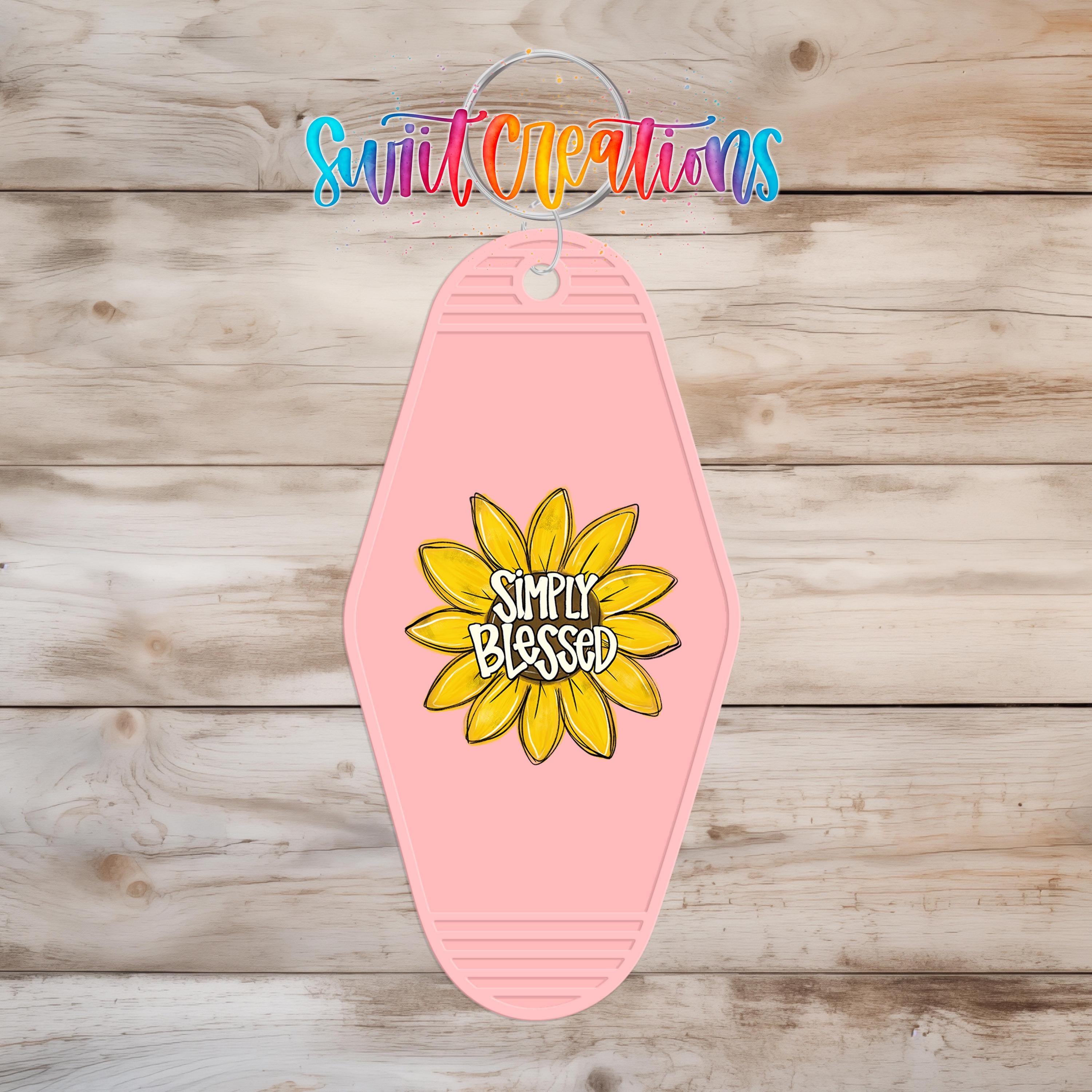 A pink keychain with a yellow flower design and the text "Simply Blessed" printed on it, hanging on a wooden surface.