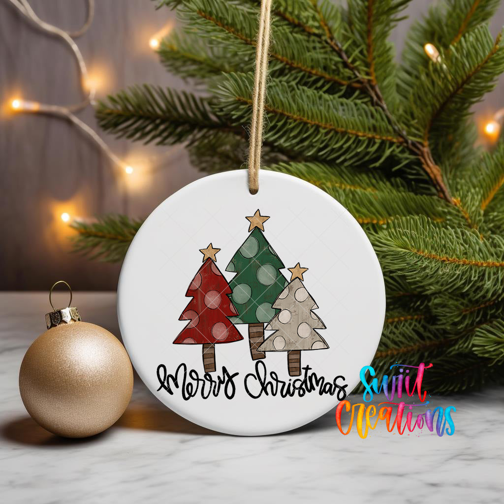 A white Christmas ornament with a design of three trees and the text "Merry Christmas" is displayed on a table, with a gold ornament and a pine tree in the background.
