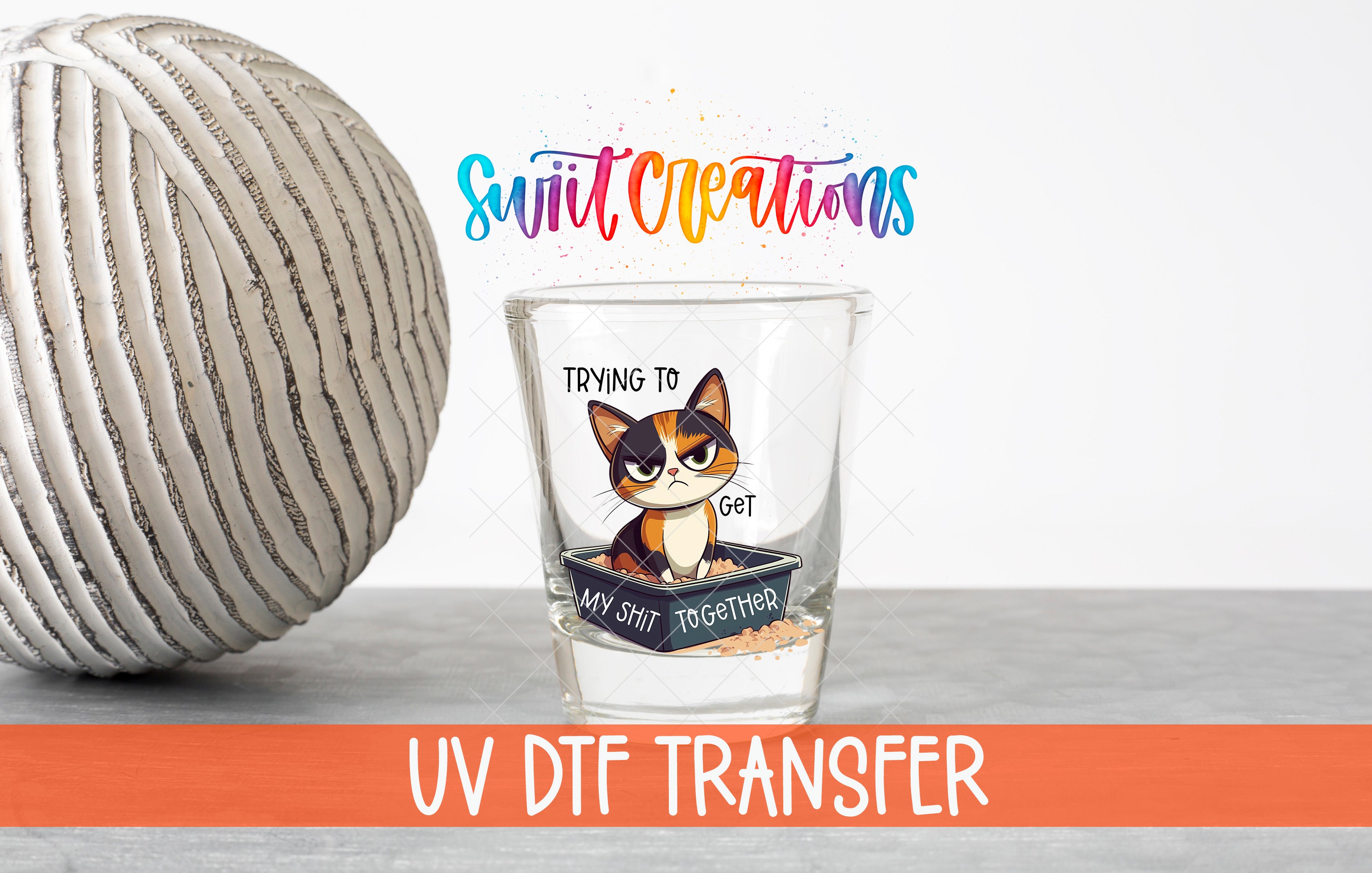 a glass with a cat illustration and text, placed next to a gray ball on a table.
