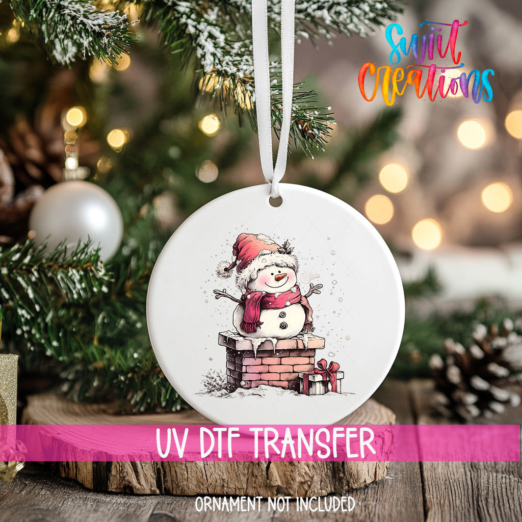 A white ornament with a snowman design hangs on a Christmas tree, surrounded by pine branches and twinkling lights.