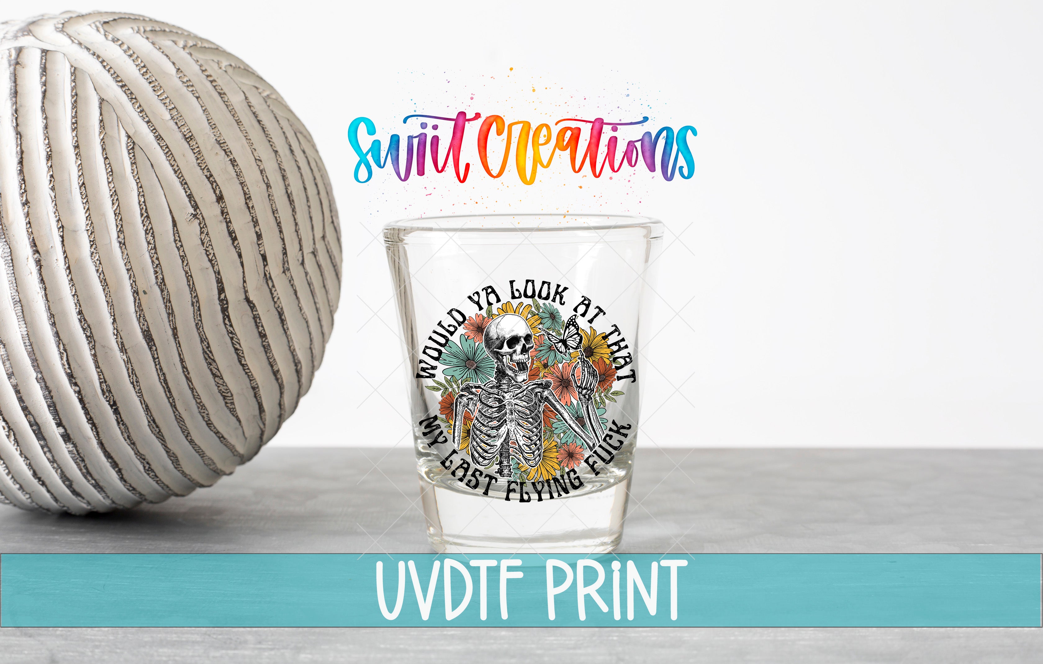 a shot glass with a skeleton design and floral elements, placed on a table next to a white ball. The text above the shot glass reads "Swift Creations" and "Uvdtf Print", and the text below the shot glass says "WOULD YA LOOK AT THAT I'D LAST FALLING FUCK".