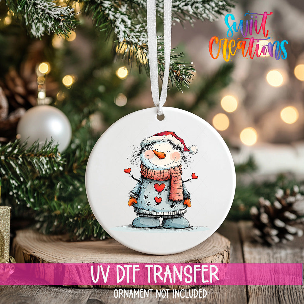 A white ceramic ornament with a snowman design hangs on a Christmas tree, surrounded by pine branches and twinkling lights.