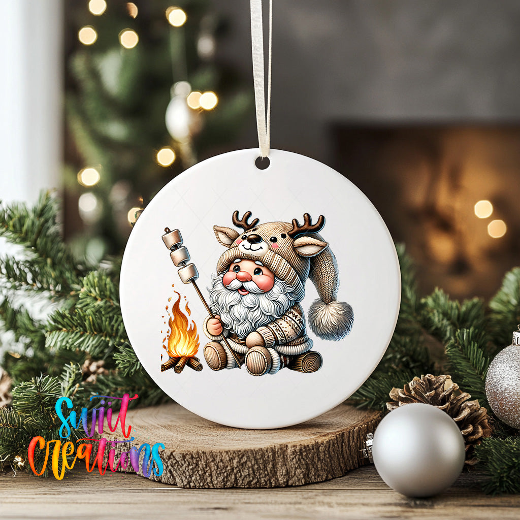 A white ornament featuring a cartoon Santa Claus character with antlers, holding a marshmallow over a campfire, surrounded by Christmas decorations and greenery.