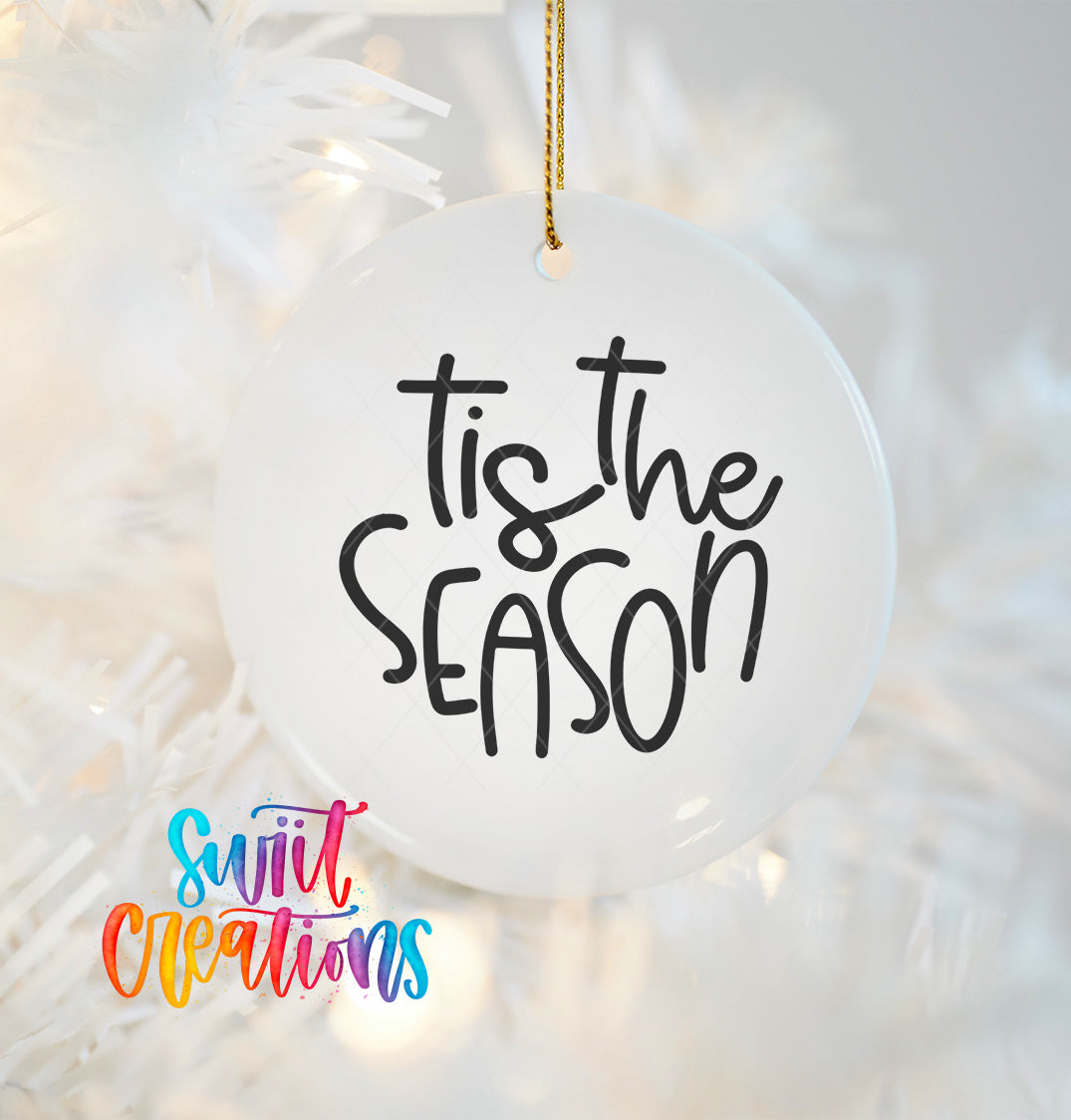 A white ornament with the text "Tis the SEASON" written on it, hanging on a Christmas tree.