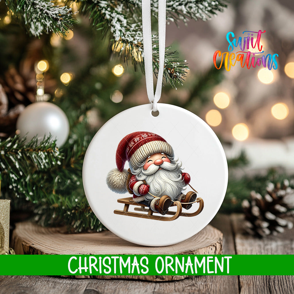 A white Christmas ornament featuring a smiling Santa Claus on a sled, hanging on a tree branch with a blurred background of lights.