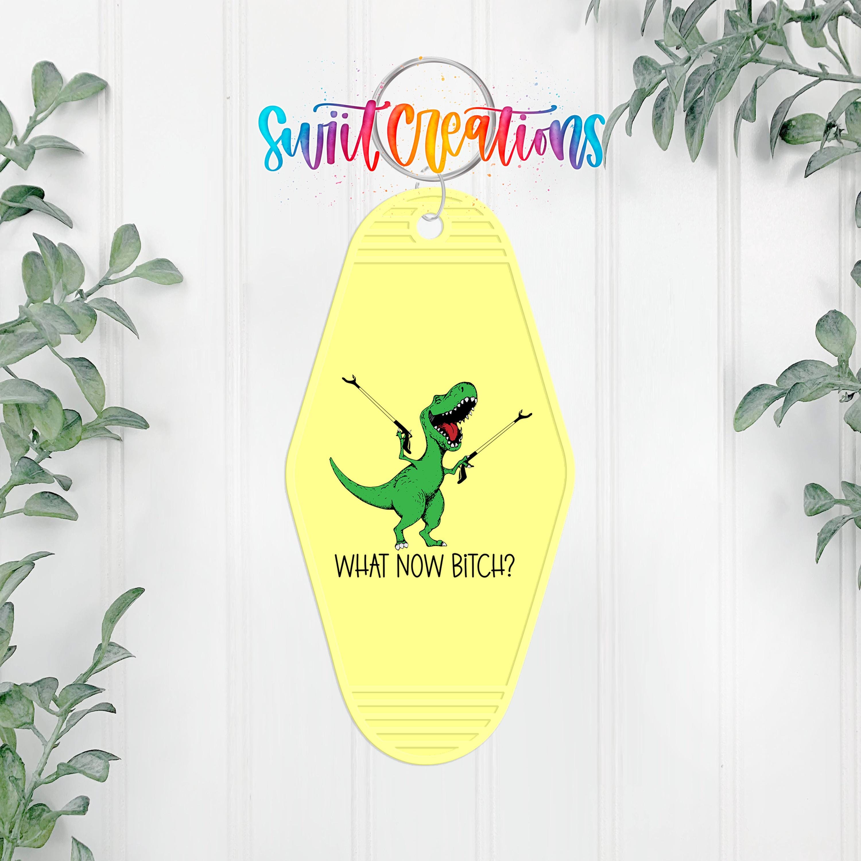 A yellow keychain with a cartoon dinosaur and the text "WHAT NOW BITCH?" hanging on a wall, surrounded by greenery.