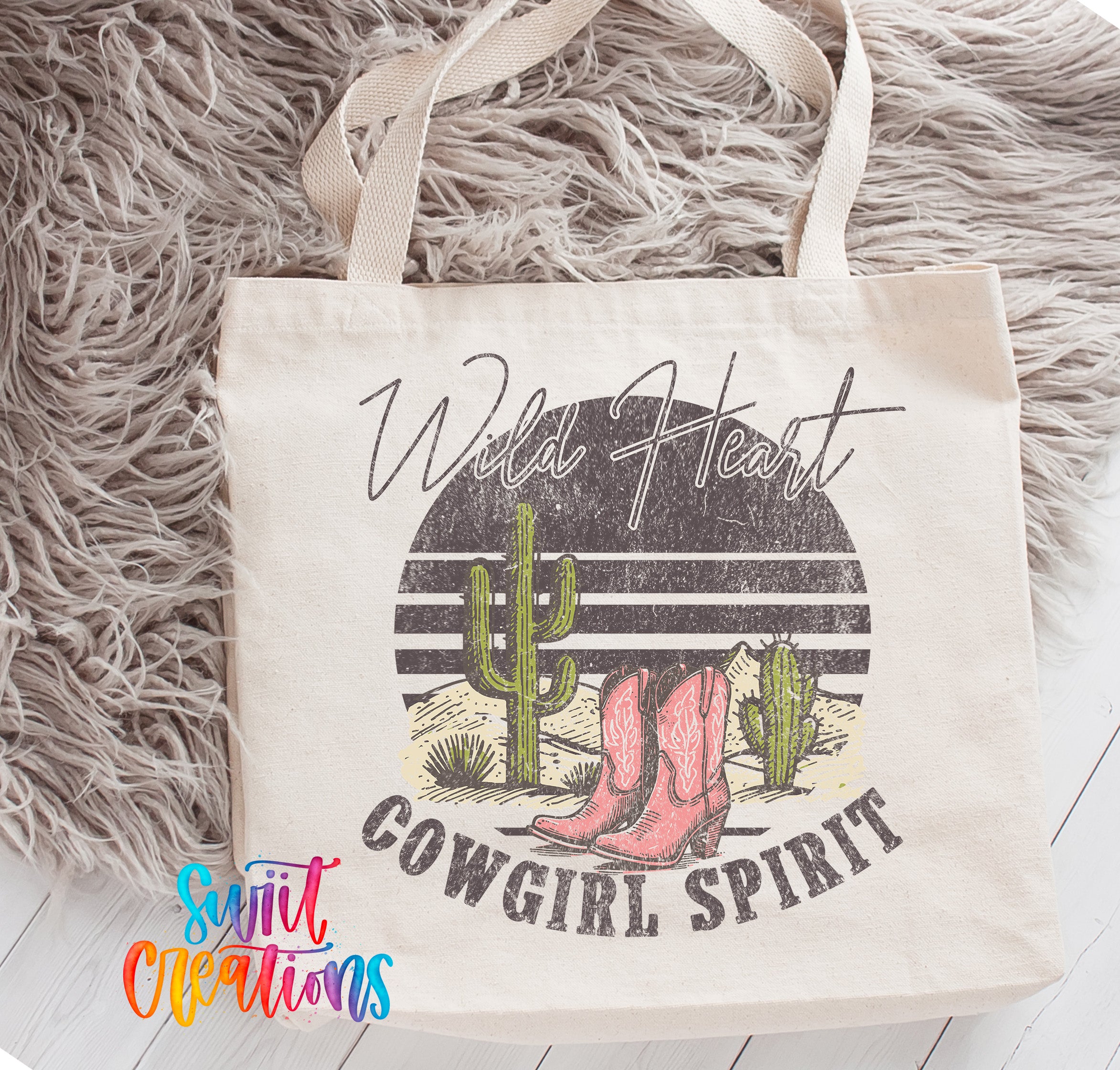 A white tote bag with a graphic design featuring a cactus, boots, and the text "Wild Heart Cowgirl Spirit" printed on it.