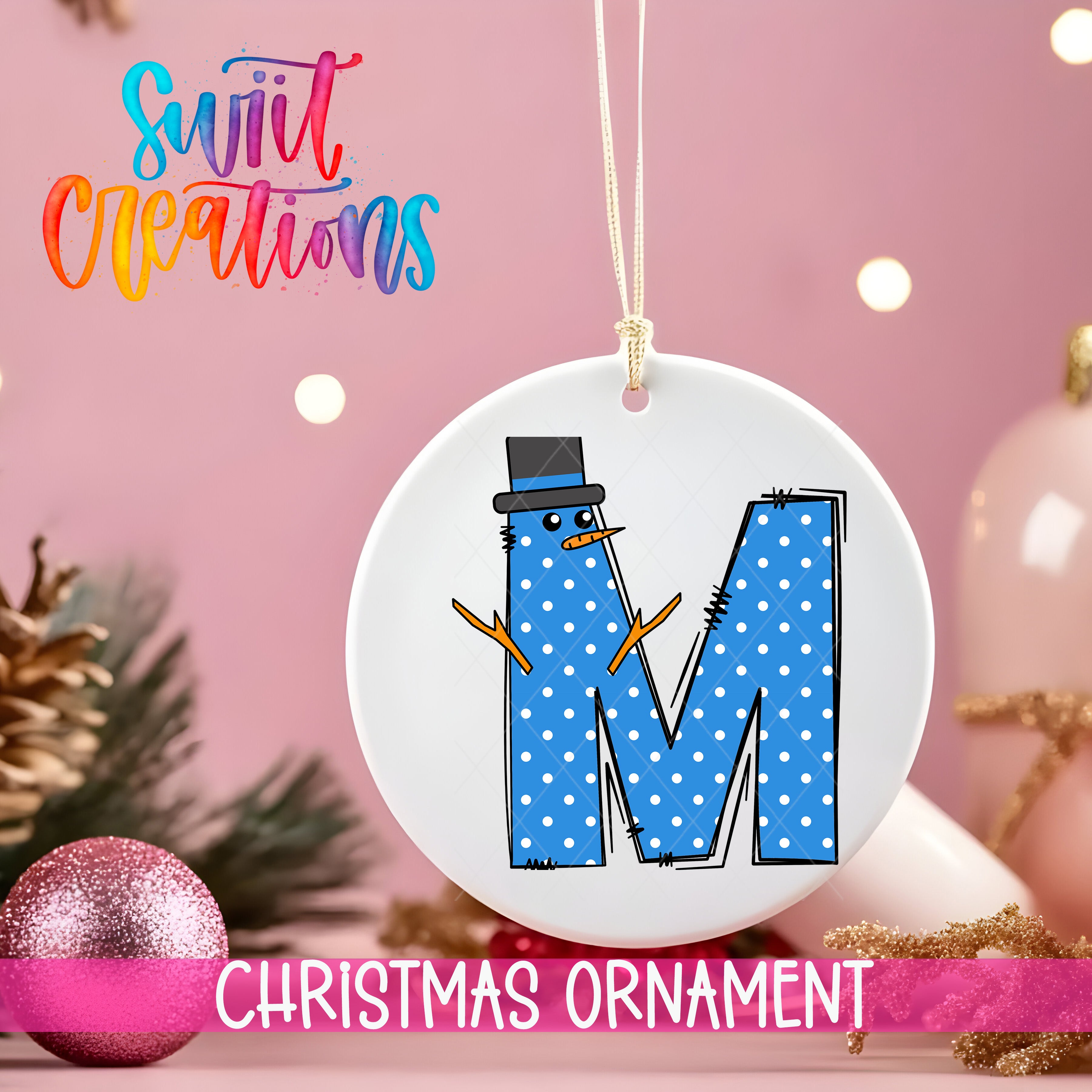 A white ornament with a blue snowman design and the letter "M" on it, hanging on a string against a pink background with Christmas decorations in the background.