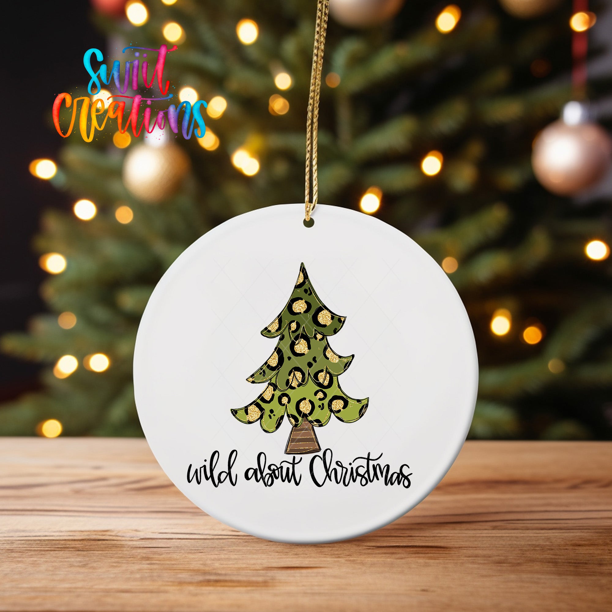 A white Christmas ornament with a leopard print tree design hangs on a wooden surface, with a blurred background of a lit Christmas tree.