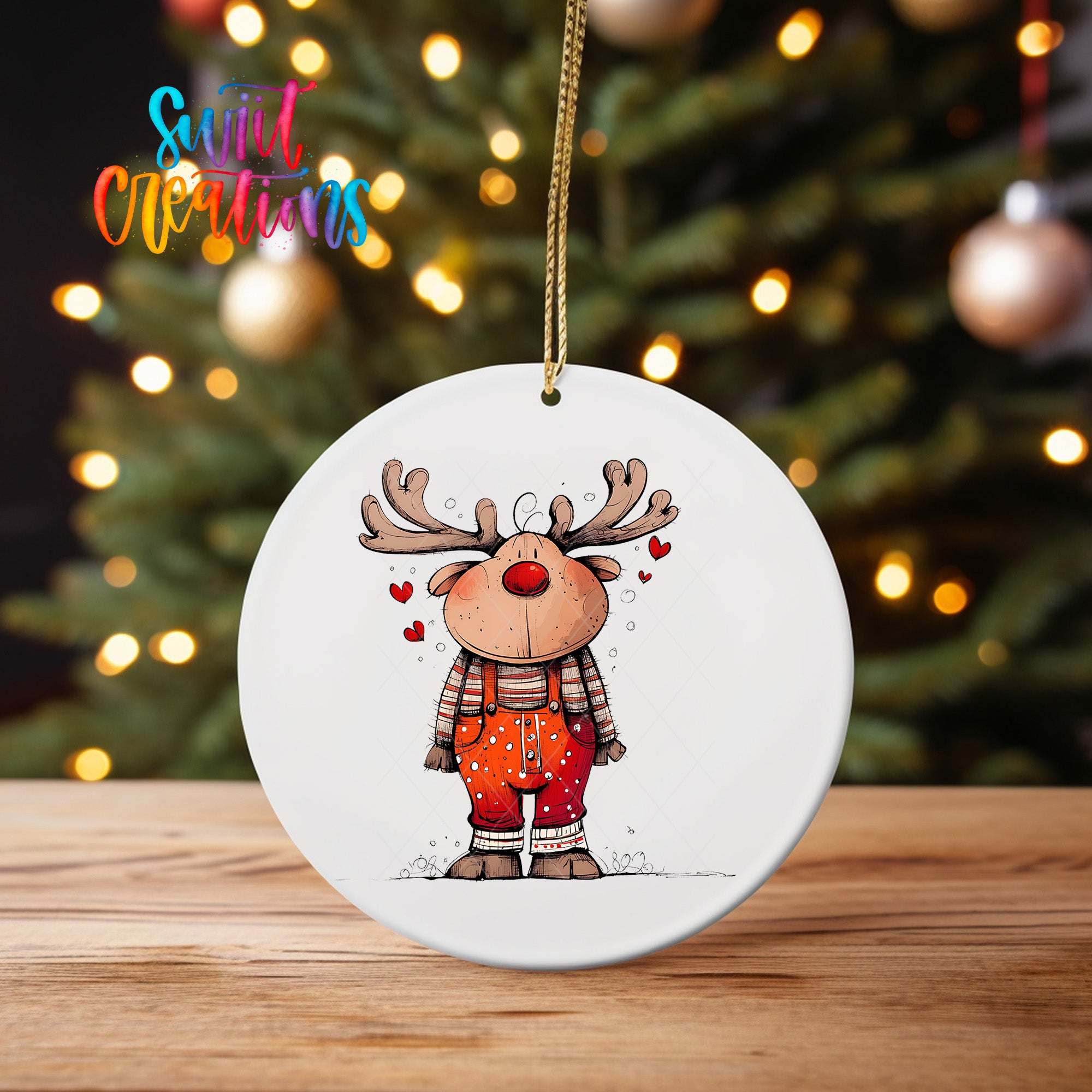 A white ornament featuring a cartoon reindeer with antlers and a red nose, standing on a wooden surface in front of a blurred Christmas tree with twinkling lights.